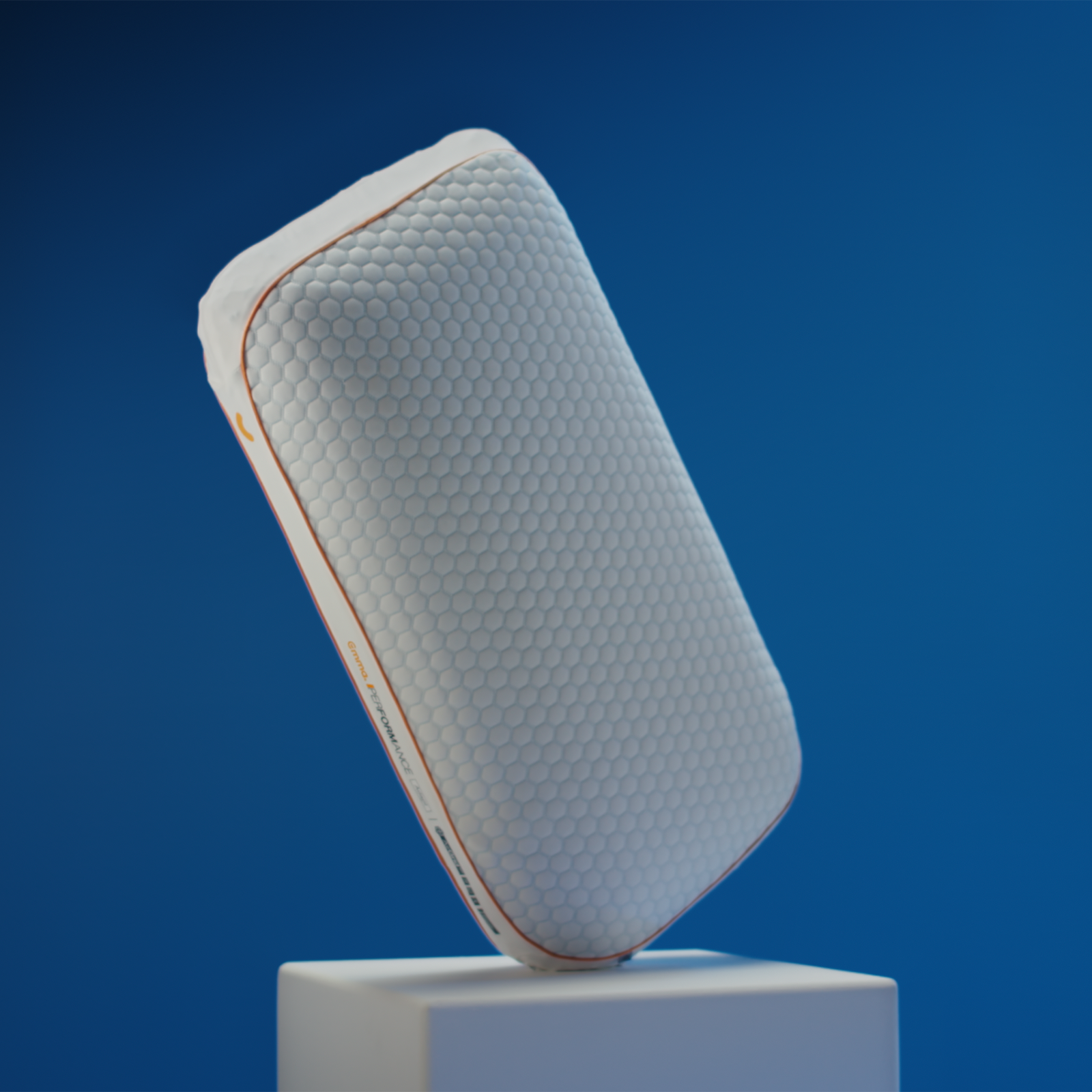 Emma Performance Adapt Pillow tilted on a white plinth showing hexagonal cover and orange trim against a blue background.