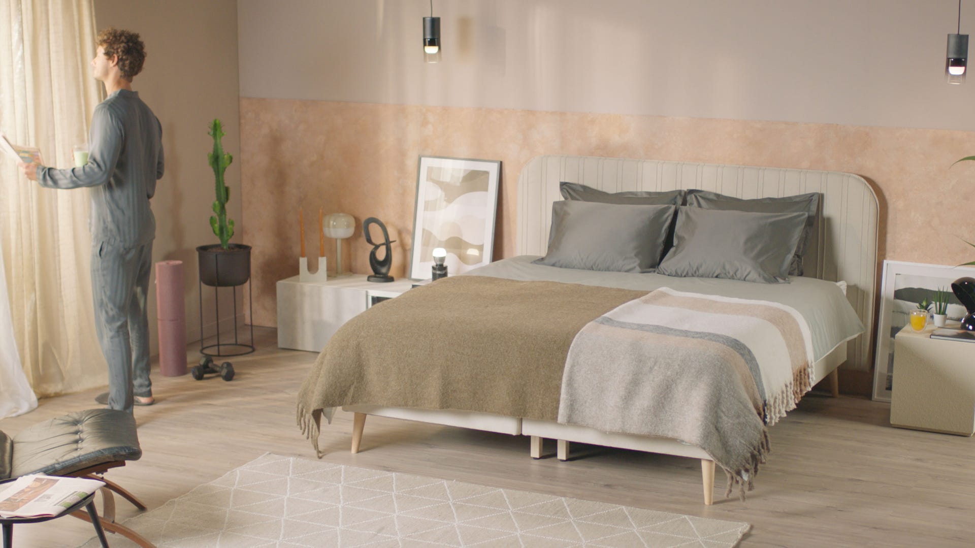 Emma Signature Boxspring in beige slaapkamer met grijs dekbed, nachtkastje met lamp en decoratie, en persoon bij het raam met boek en kopje.