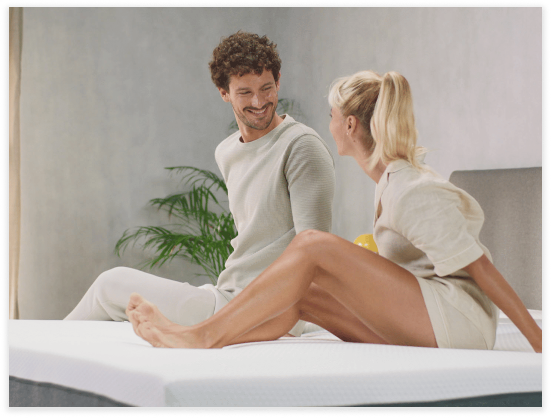 Un homme et une femme sont assis sur un lit, se souriant l'un à l'autre dans une chambre à coucher moderne et lumineuse avec une plante en arrière-plan. Tous deux sont habillés de façon décontractée et semblent détendus et heureux.