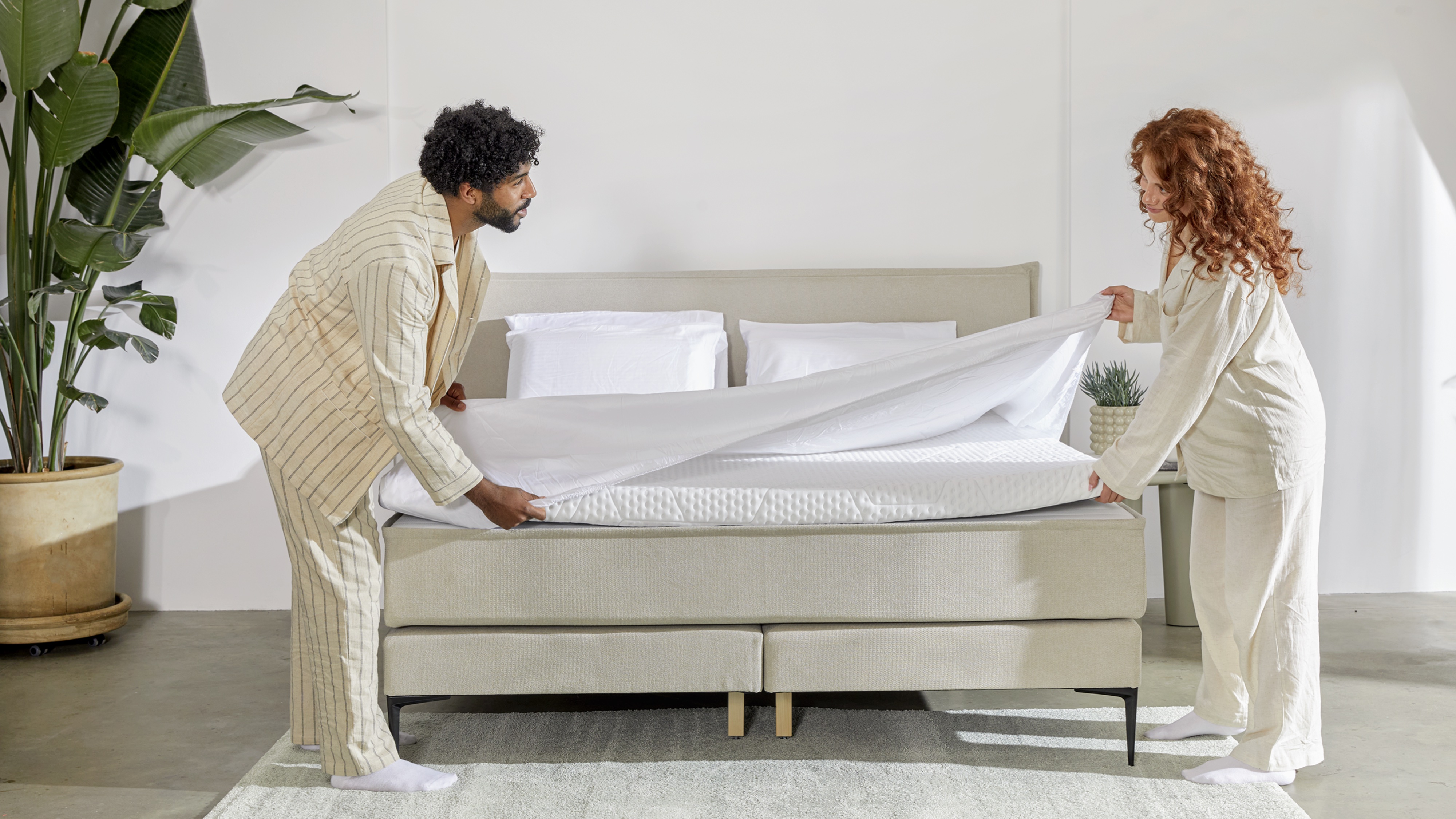 Twee personen maken een Emma Signature Boxspring op met wit hoeslaken; bed met beige gestoffeerd frame, drie witte kussens en grote groene kamerplant links, bijzettafeltje met plant rechts; lichte slaapkamer met minimalistische inrichting en zacht vloerkleed.