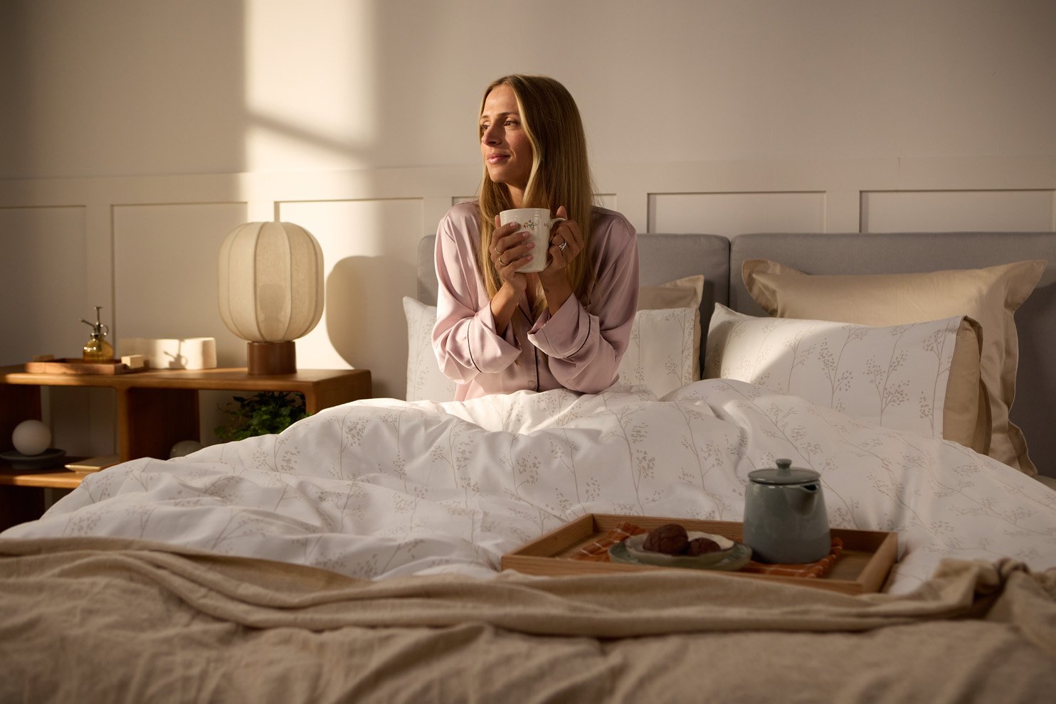 Persoon die rechtop in bed zit met een mok in de handen, omringd door licht beddengoed met een subtiel botanisch patroon en een houten nachtkastje met lamp en decoratie naast het bed.
