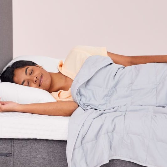 Persoon slaapt op een bed met een lichtgrijze Emma Verzwaringsdeken, witte kussens en een grijs gestoffeerd hoofdeinde – straalt rust en comfort uit.