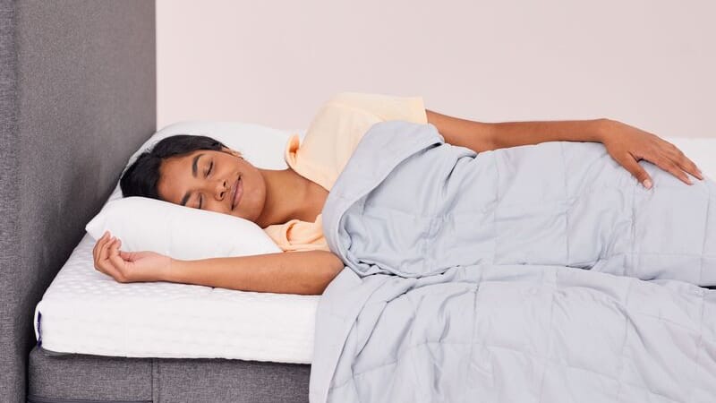 Persoon slaapt op een bed met een lichtgrijze Emma Verzwaringsdeken, witte kussens en een grijs gestoffeerd hoofdeinde – straalt rust en comfort uit.