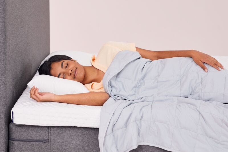 Persoon slaapt op een bed met een lichtgrijze Emma Hug Verzwaringsdeken, witte kussens en een grijs gestoffeerd hoofdeinde – straalt rust en comfort uit.