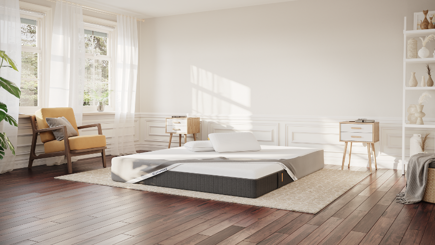 Minimalistische slaapkamer met een Emma Hybrid Premium matras op een lichtgrijs tapijt, deels bedekt met een grijze matrasbeschermer. Op het matras liggen twee witte Emma Diamond Kussens met zwarte rand. Rechts staat een zwart bijzettafeltje met houten poten, waarop een zwarte vaas met decoratieve takjes en een tijdschrift liggen. Deze afbeelding vertegenwoordigt de Emma Hybrid Premium Deal met gratis matrasbeschermer, Wolkenkussen(s) en Hybrid Premium matras.