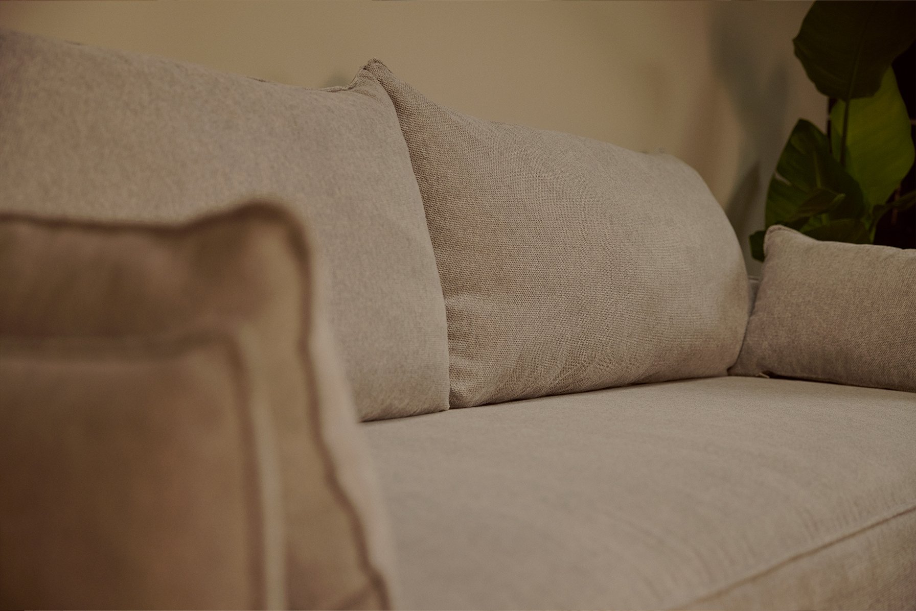 Canapé convertible gris clair déplié en position lit, posé sur un tapis beige dans un salon lumineux. À gauche, grande fenêtre avec rideaux blancs laissant entrer la lumière naturelle. Sous la fenêtre, console en bois avec lampe, objets déco et paniers tressés. À droite, meuble en bois avec livres, vases et tableau abstrait encadré. Suspension ronde en fibres naturelles au plafond. Moulures en bois au sol et au plafond renforçant l’ambiance cosy et épurée.