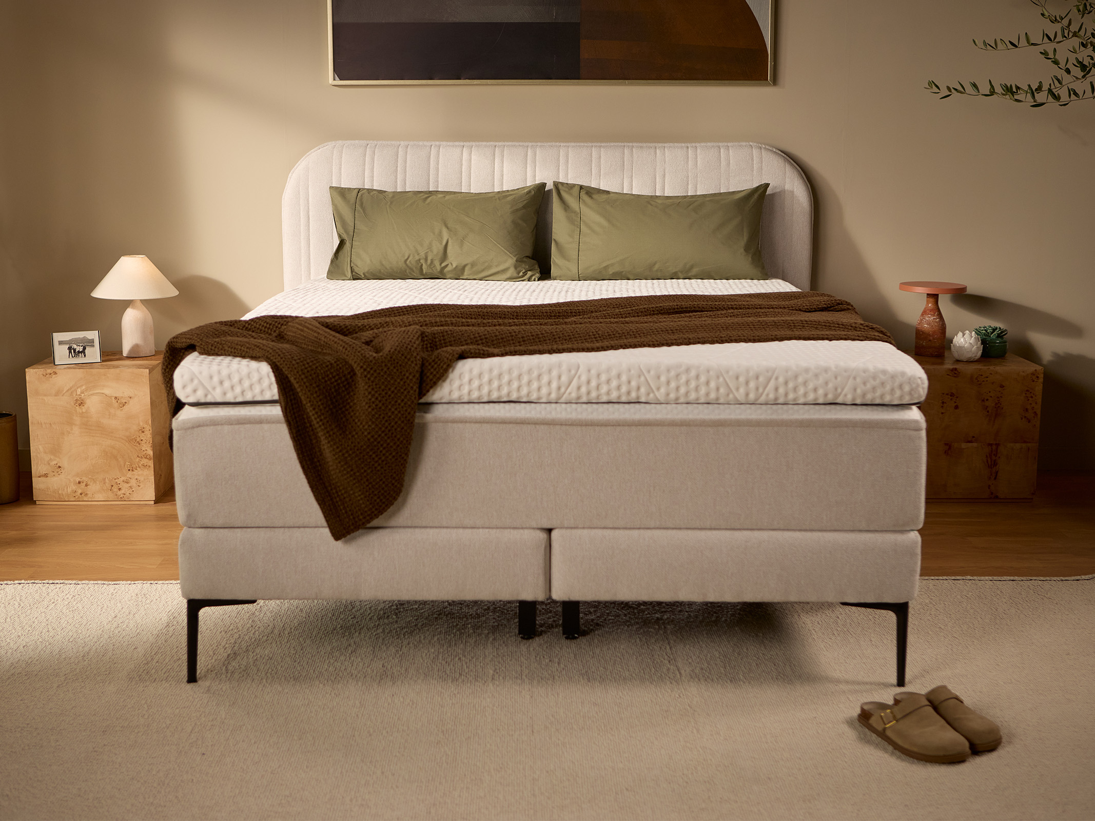 Light beige upholstered box spring bed with channel-stitched headboard, pillows, and bedding, shown from the front.