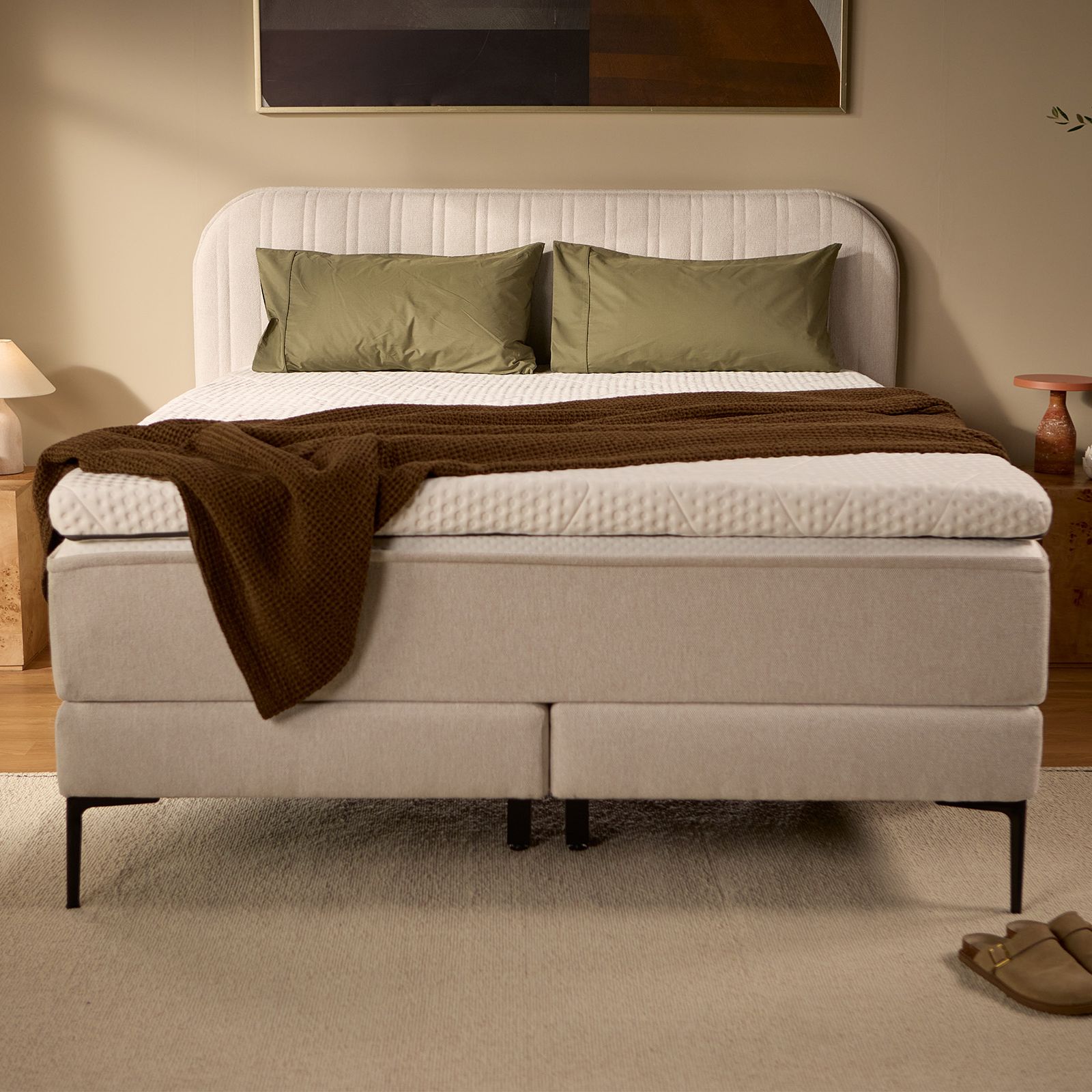 Light beige upholstered box spring bed with channel-stitched headboard, pillows, and bedding, shown from the front.