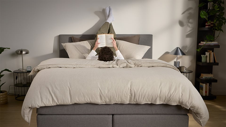 Grey upholstered bed with white mattress and beige bedding, shown with a person reading.
