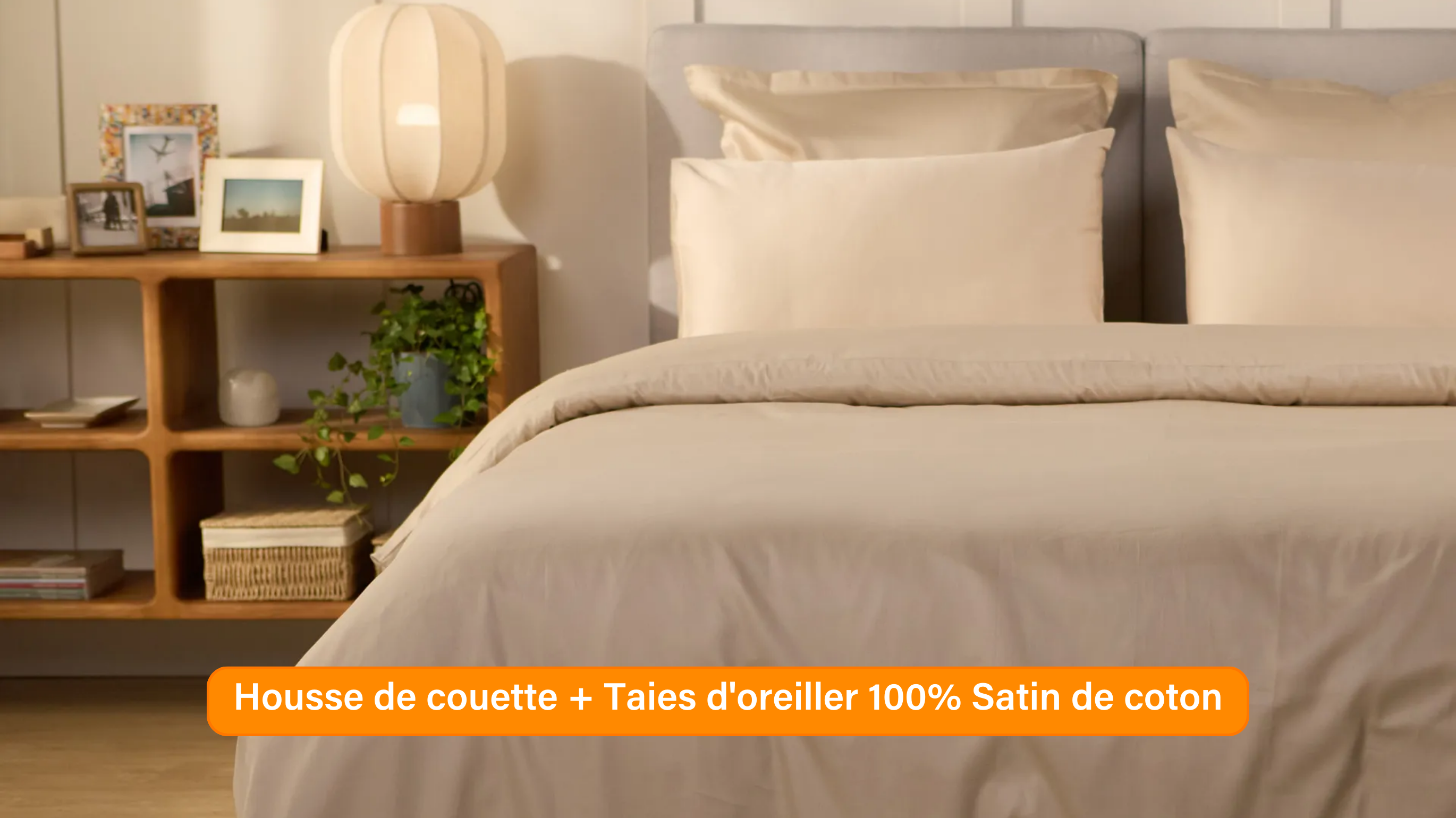 Chambre élégante avec lit soigneusement préparé, habillé d’une parure de lit 100 % satin de coton de couleur blanche ou beige clair. Plusieurs oreillers sont disposés contre un mur à panneaux. De chaque côté du lit, des tables de chevet avec lampe et objets décoratifs. Un banc rose est placé au pied du lit, assorti à un tabouret près de la table de chevet. Le sol en bois clair et les murs neutres créent une ambiance douce et raffinée.