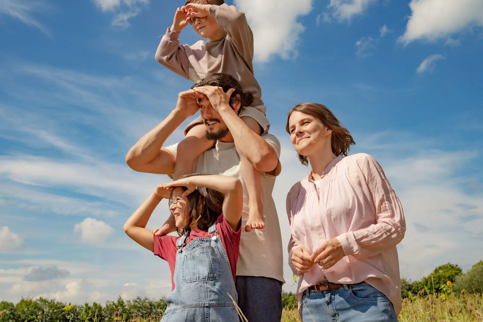 Familia con dos adultos y dos niños al aire libre en un día soleado, todos mirando al horizonte con la mano sobre los ojos, sugiriendo contemplación o curiosidad. El hombre lleva a un niño sobre los hombros, mientras la mujer y otro niño observan al lado. El paisaje natural incluye vegetación verde, cielo azul con nubes y una atmósfera ligera y relajada.