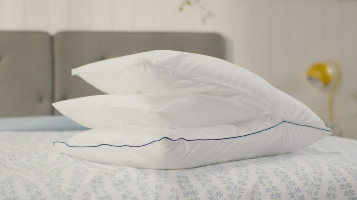 Original Cooling Comfort Kussen op bed om alle verwijderbare lagen te zien. Op de achtergrond staat een grijs gestoffeerd hoofdbord en een gele lamp op een nachtkastje. Deze afbeelding toont het luchtige en stijlvolle ontwerp van het Emma Wolkenkussen in een huiselijke setting.