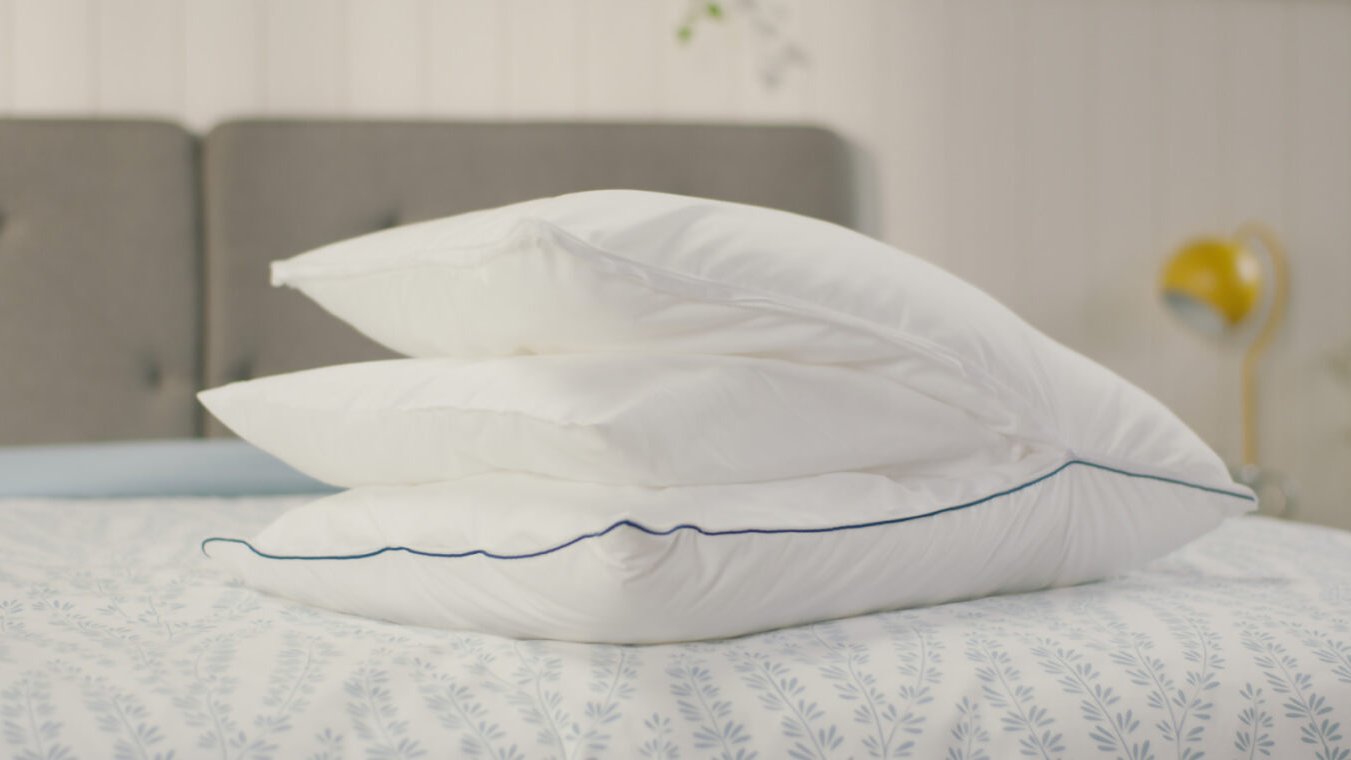 Original Cooling Comfort Kussen op bed om alle verwijderbare lagen te zien. Op de achtergrond staat een grijs gestoffeerd hoofdbord en een gele lamp op een nachtkastje. Deze afbeelding toont het luchtige en stijlvolle ontwerp van het Emma Wolkenkussen in een huiselijke setting.