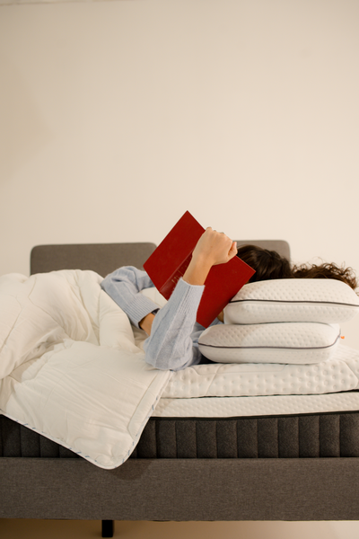 Femme allongée sur le côté dans un lit, la tête reposant sur deux oreillers, lisant un livre.