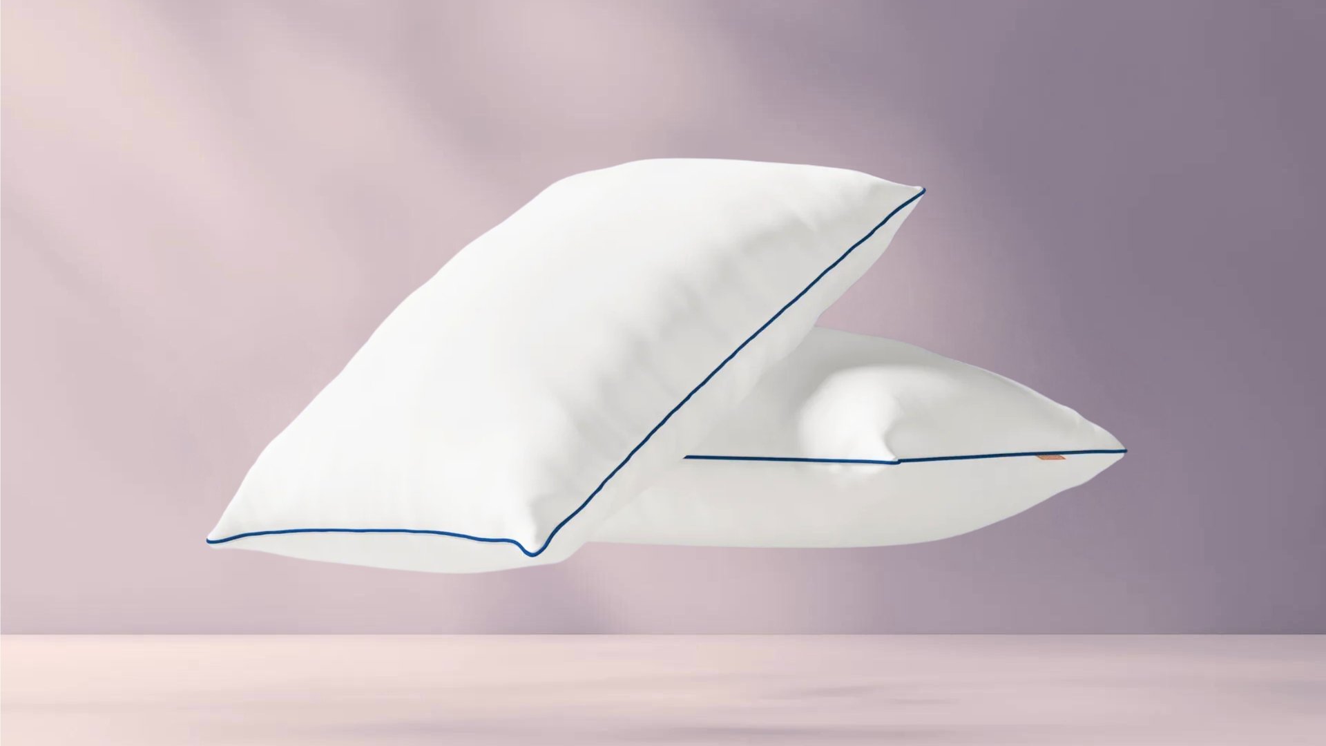 Twee witte kussens met donkerblauwe bies, waarbij één kussen schuin tegen het andere leunt, tegen een zachtroze achtergrond.