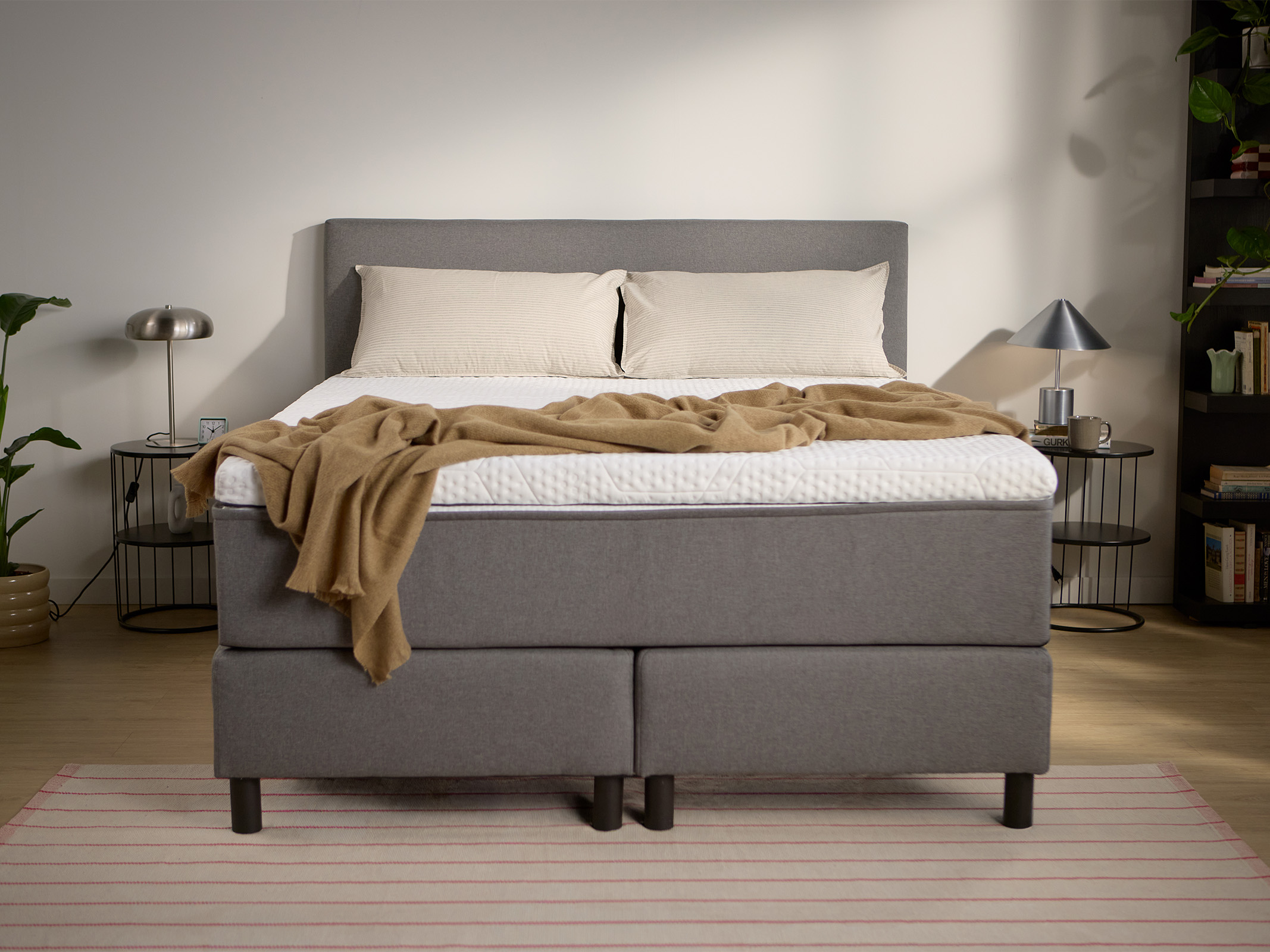 Dark grey upholstered box spring bed with headboard, visible central support legs, and bedding, shown from the front.