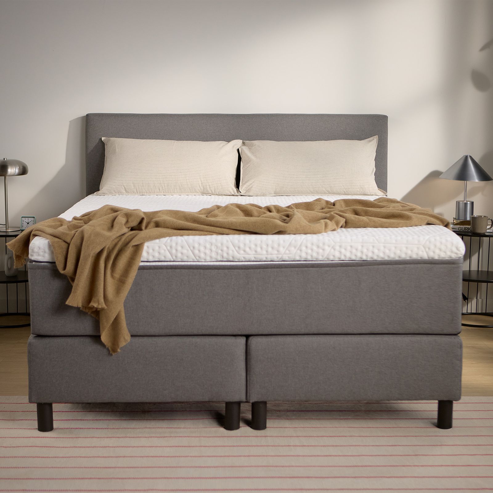Dark grey upholstered box spring bed with headboard, visible central support legs, and bedding, shown from the front.