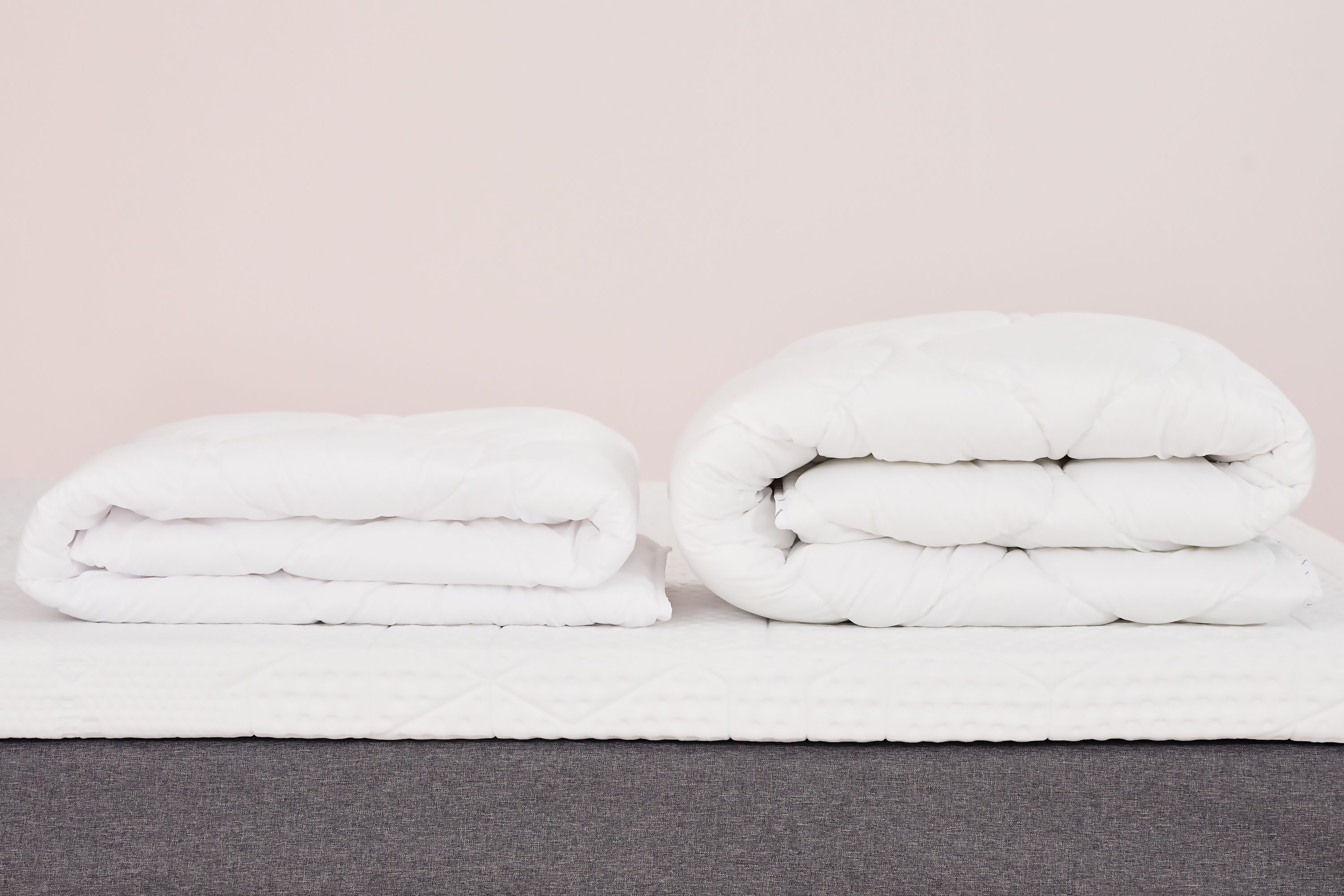 Due piumoni bianchi piegati su letto con base grigia e materasso bianco, uno compatto e uno arrotolato