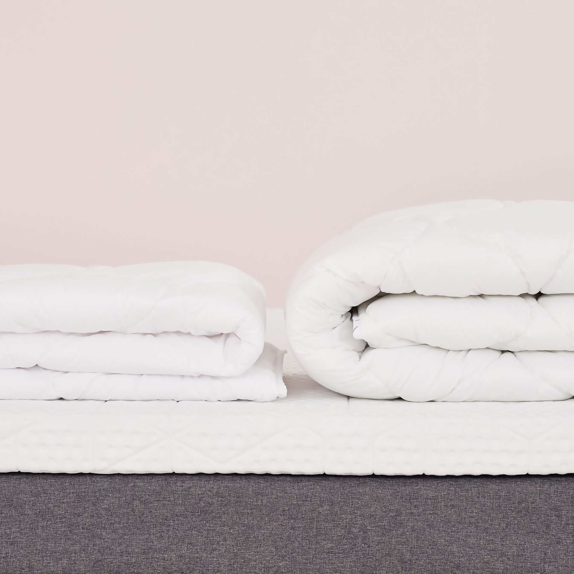 Due piumoni bianchi piegati su letto con base grigia e materasso bianco, uno compatto e uno arrotolato