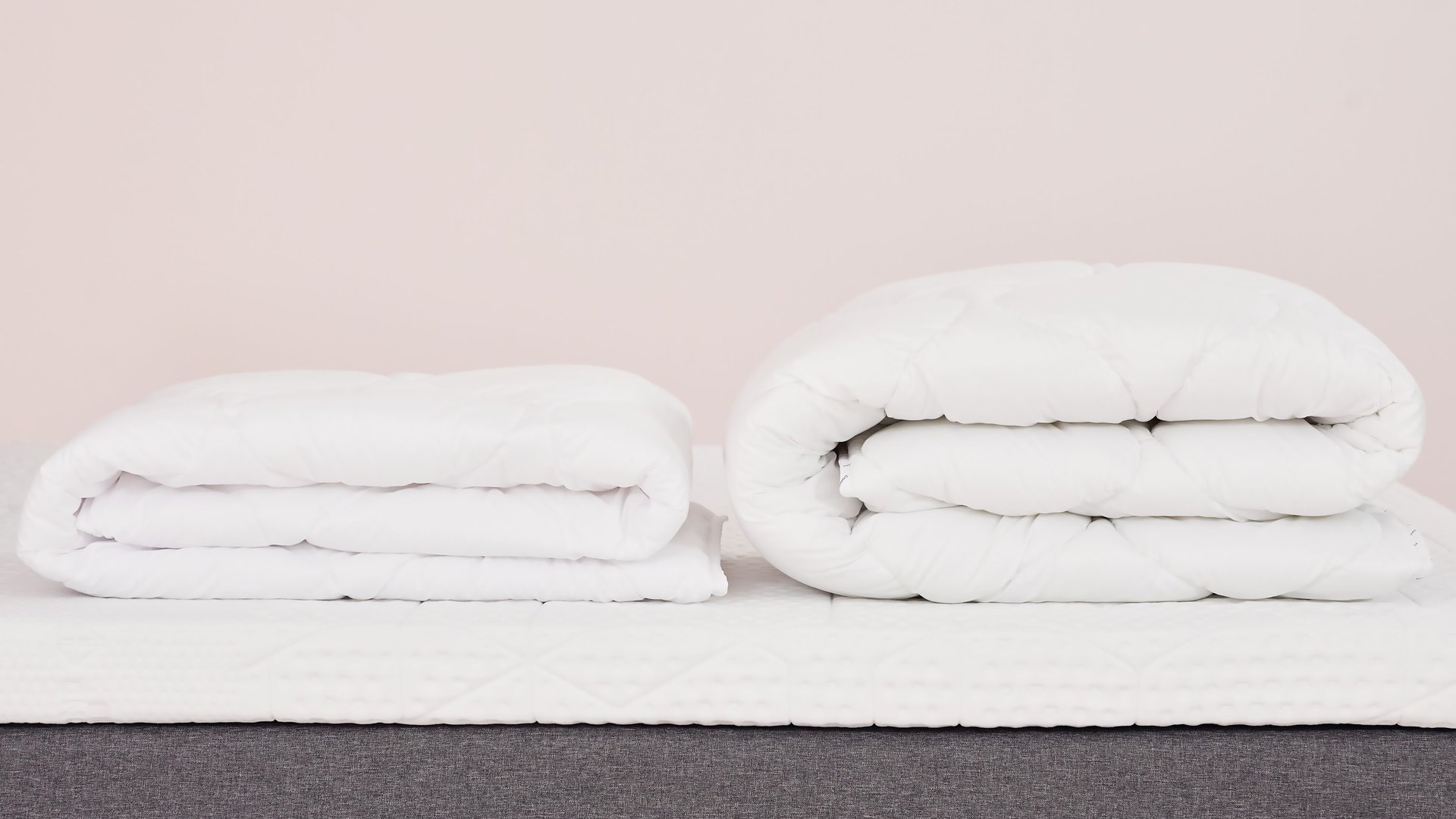 Due piumoni bianchi piegati su letto con base grigia e materasso bianco, uno compatto e uno arrotolato