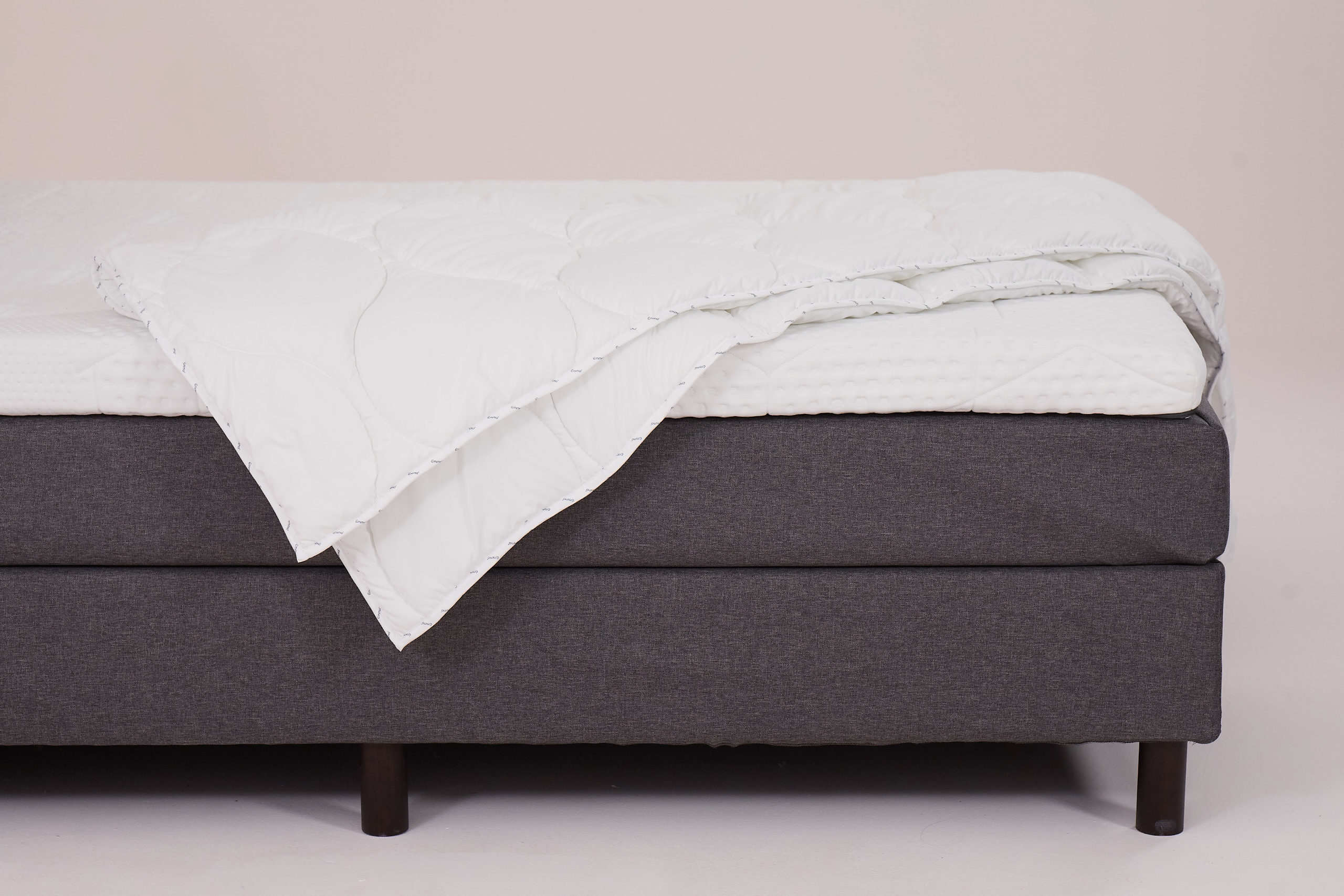 Letto con base grigia, gambe in legno scuro, coprimaterasso trapuntato bianco e piumone piegato.