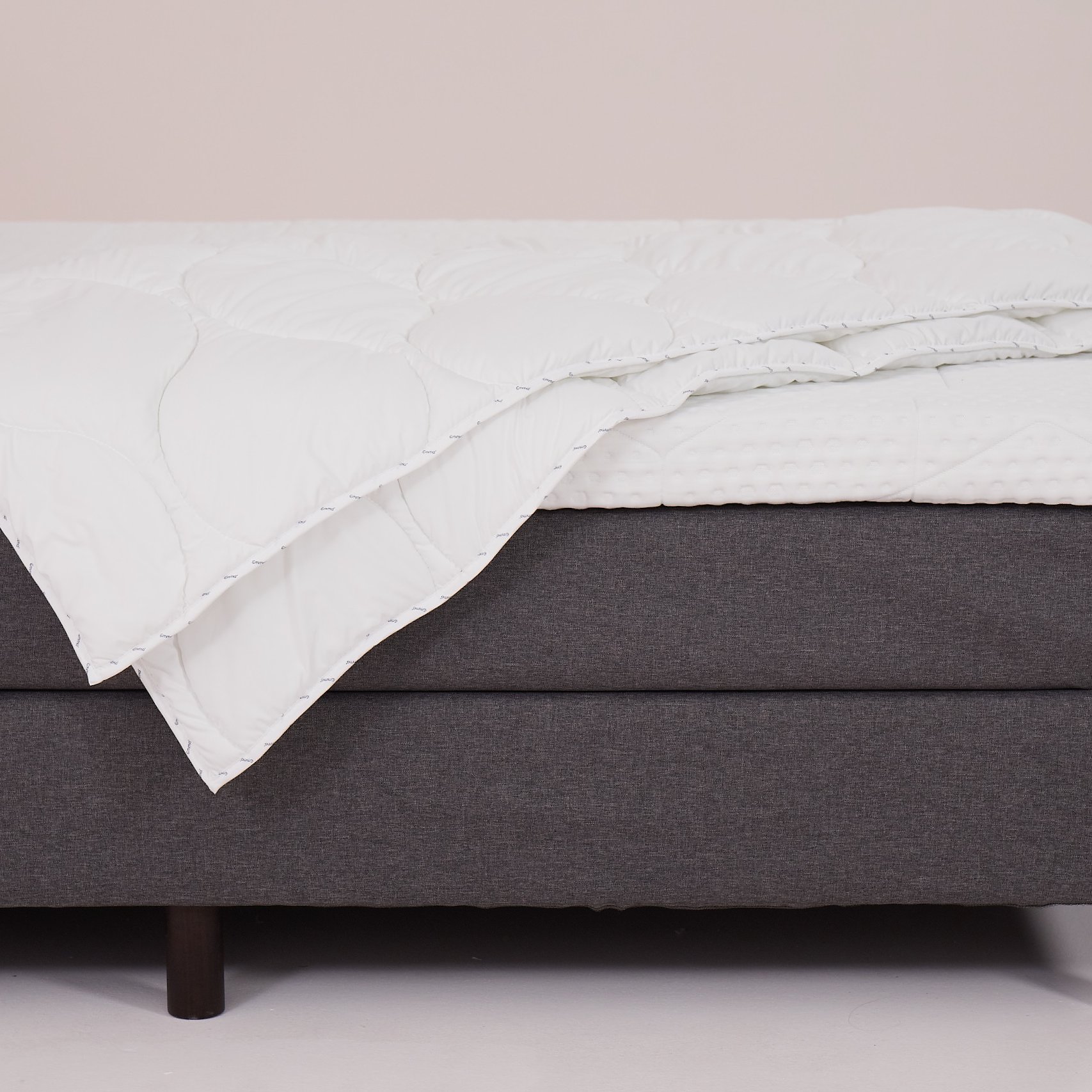 Letto con base grigia, gambe in legno scuro, coprimaterasso trapuntato bianco e piumone piegato.