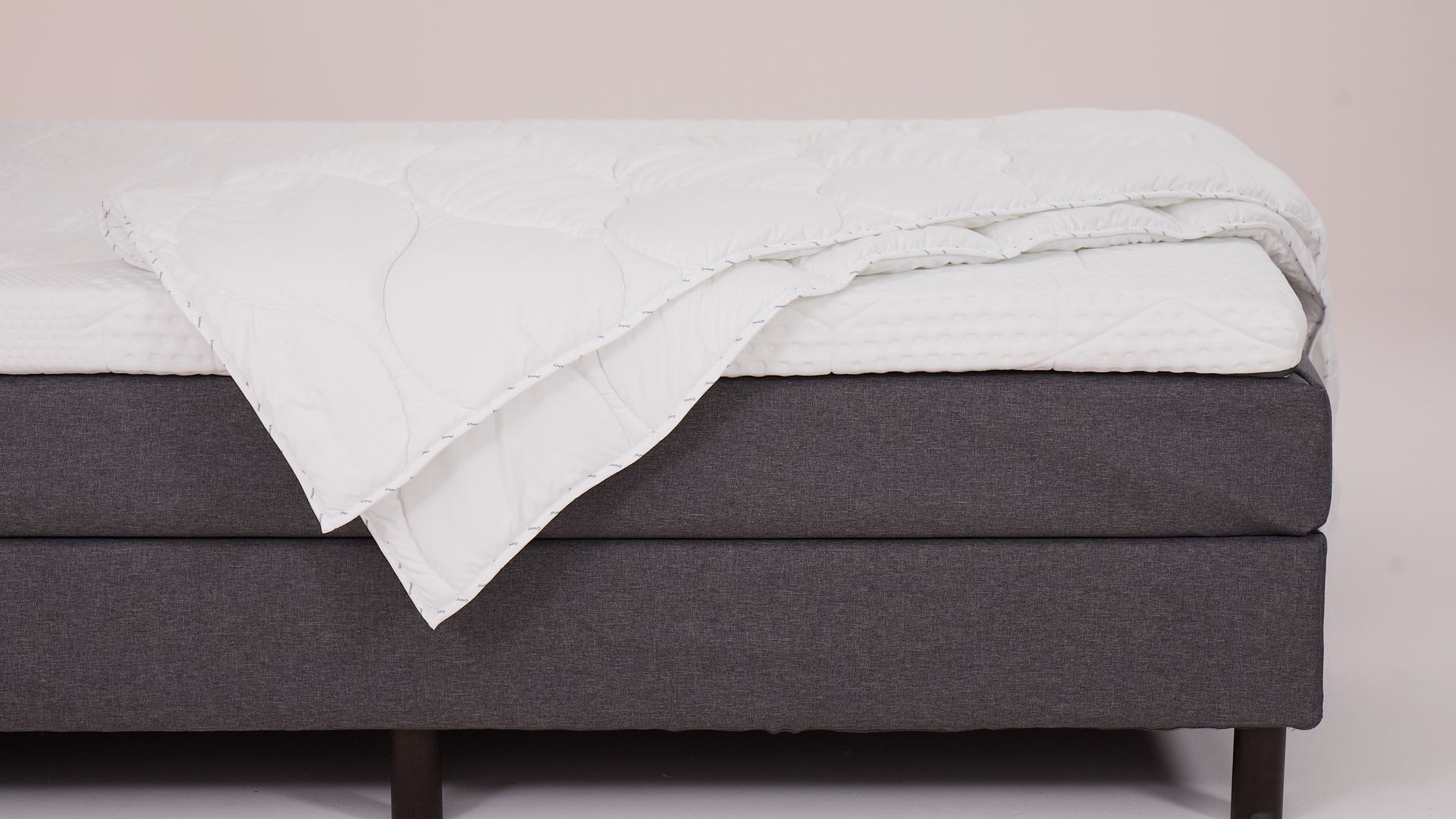 Letto con base grigia, gambe in legno scuro, coprimaterasso trapuntato bianco e piumone piegato.
