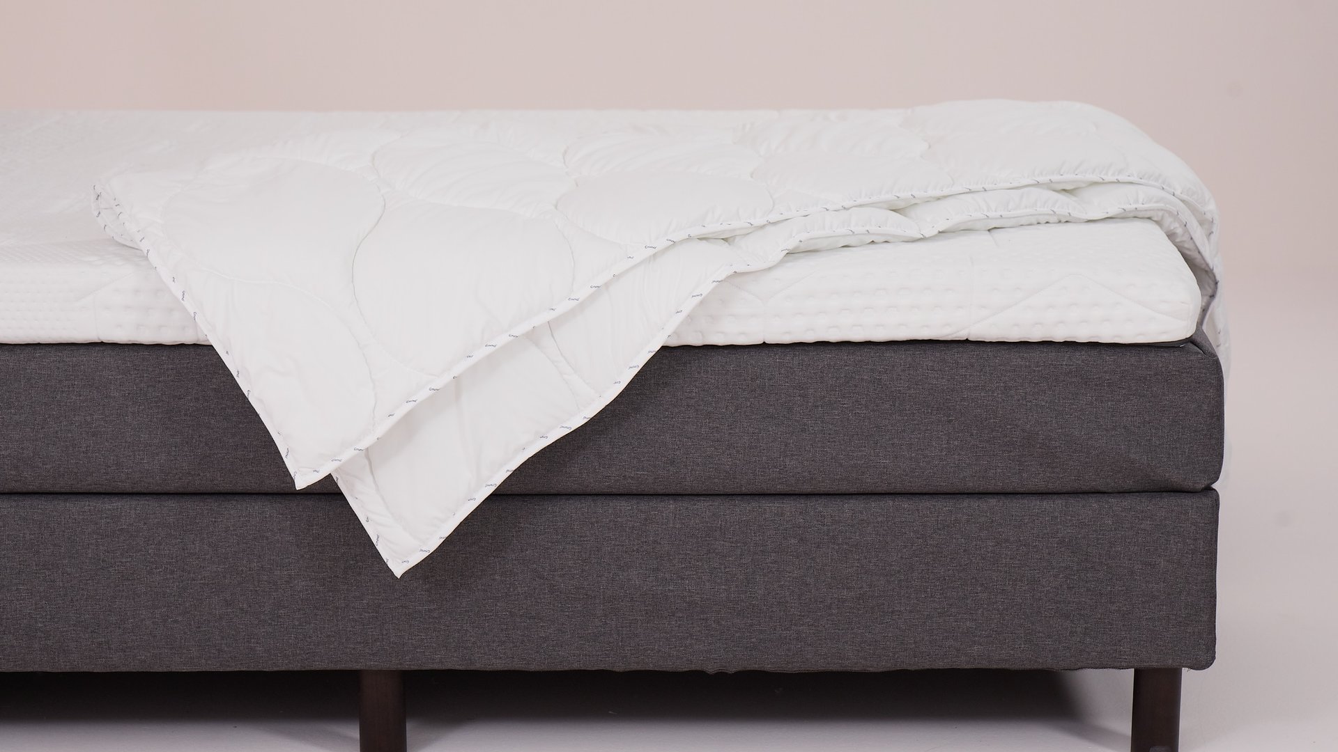 Letto con base grigia, gambe in legno scuro, coprimaterasso trapuntato bianco e piumone piegato.