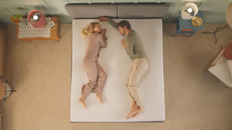 Twee mensen liggen ontspannen op een bed in een slaapkamer met warme verlichting. De kamer bevat een nachtkastje met een lamp en een boek, een tafel met een stoel, en een kast met een plant en een kaars.