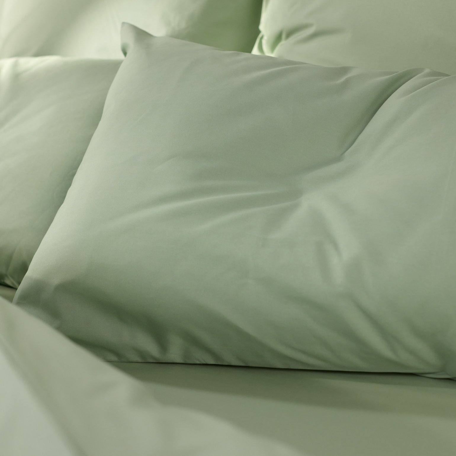 Close‑up van een opgemaakt bed met lichtgroen beddengoed en meerdere kussens in dezelfde zachte groentint.