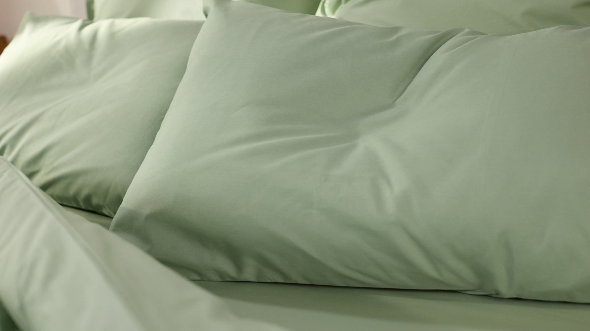 Close‑up van een opgemaakt bed met lichtgroen beddengoed en meerdere kussens in dezelfde zachte groentint.