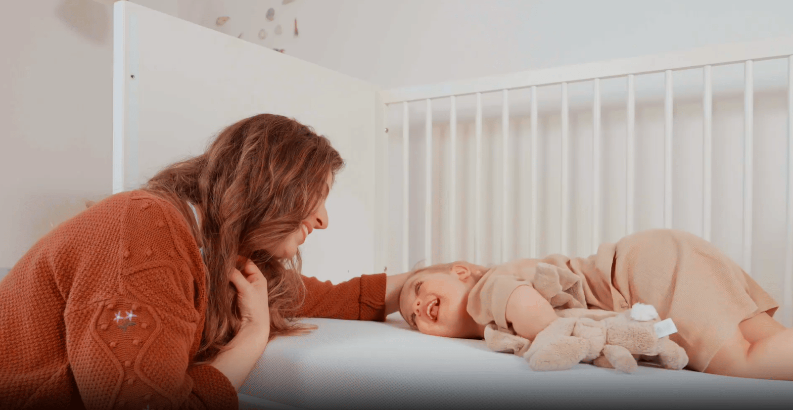 Volwassene leunt tegen een kinderledikant terwijl een kind op de Emma Baby & Kindermatras ligt met een pluche knuffel.