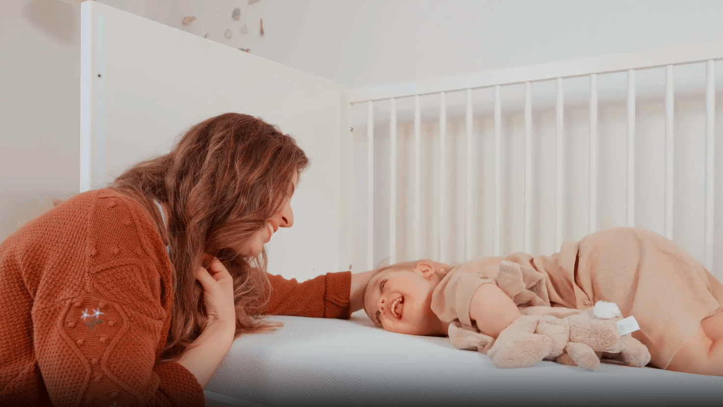 Volwassene leunt tegen een kinderledikant terwijl een kind op de Emma Baby & Kindermatras ligt met een pluche knuffel.