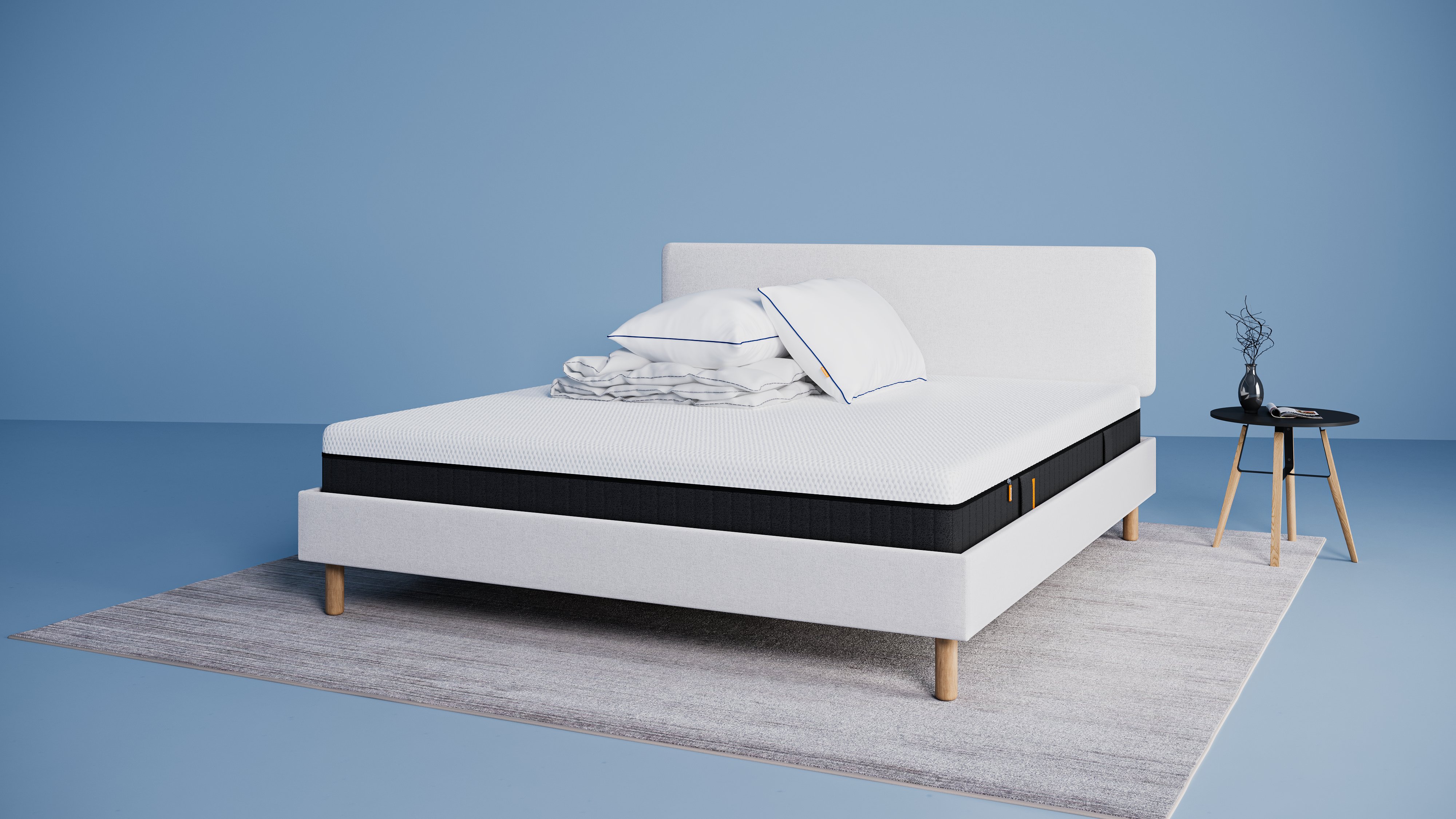 Modern bed met lichtgrijs stoffen frame en matras, opgemaakt met witte kussens en dekbed, naast een kleine zwarte ronde bijzettafel met vaas op een licht vloerkleed tegen een blauwe achtergrond.