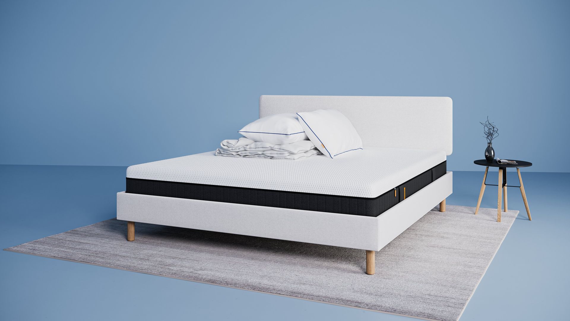 Modern bed met lichtgrijs stoffen frame en matras, opgemaakt met witte kussens en dekbed, naast een kleine zwarte ronde bijzettafel met vaas op een licht vloerkleed tegen een blauwe achtergrond.