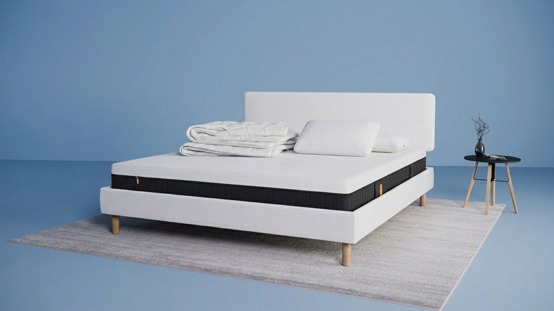 Modern bed met lichtgrijs stoffen frame en matras, opgemaakt met witte kussens en dekbed, naast een kleine zwarte ronde bijzettafel met vaas op een licht vloerkleed tegen een blauwe achtergrond.