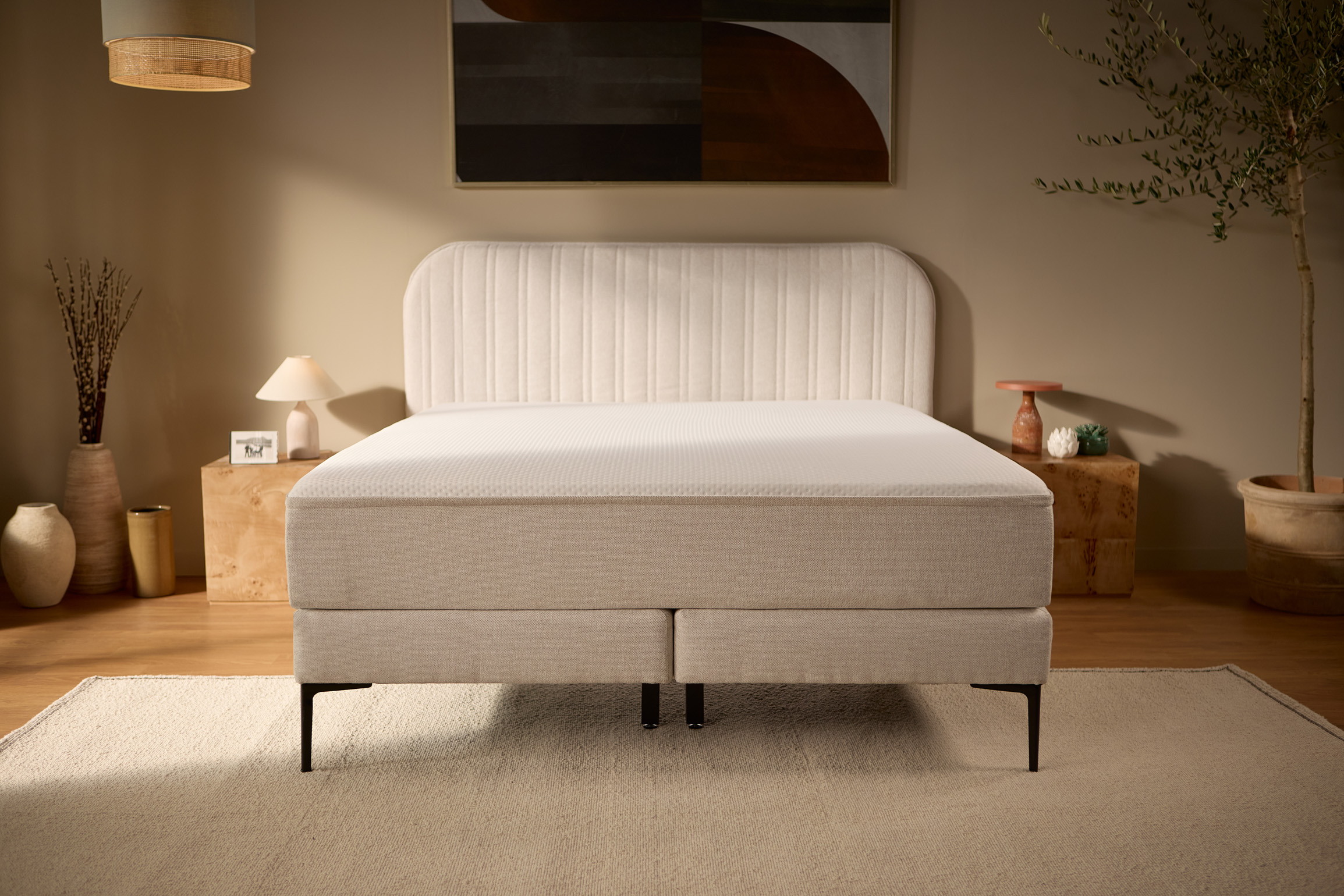 Vooraanzicht van het volledige beige bed met een gestikt hoofdbord, een verhoogde boxspringbasis, slanke buitenpoten en stevigere centrale ondersteuningspoten.