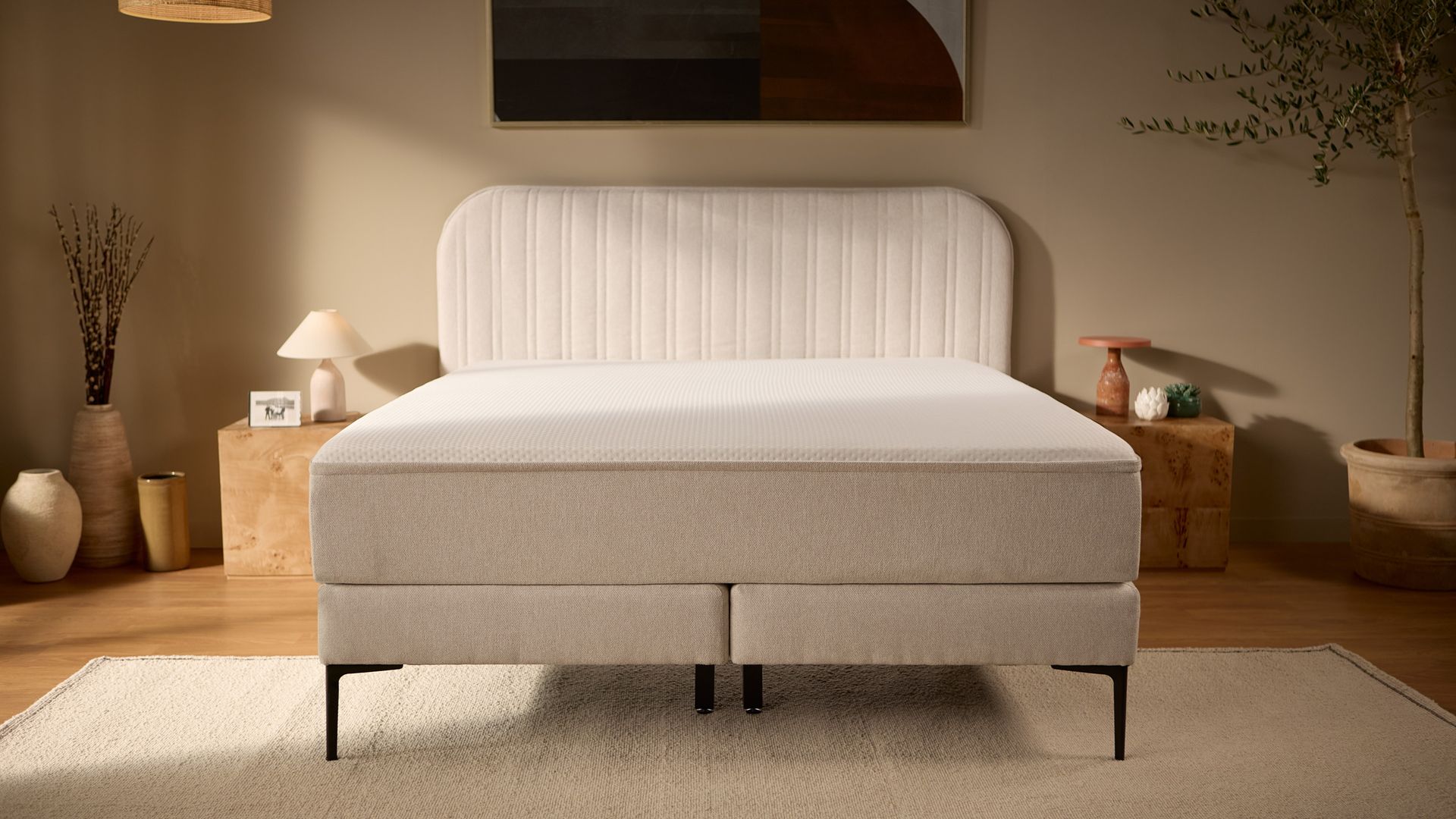 Frontansicht des kompletten beigen Bettes mit gestepptem Kopfteil, erhöhtem Boxspring-Unterbau, schlanken Außenbeinen und robusteren mittigen Stützbeinen.