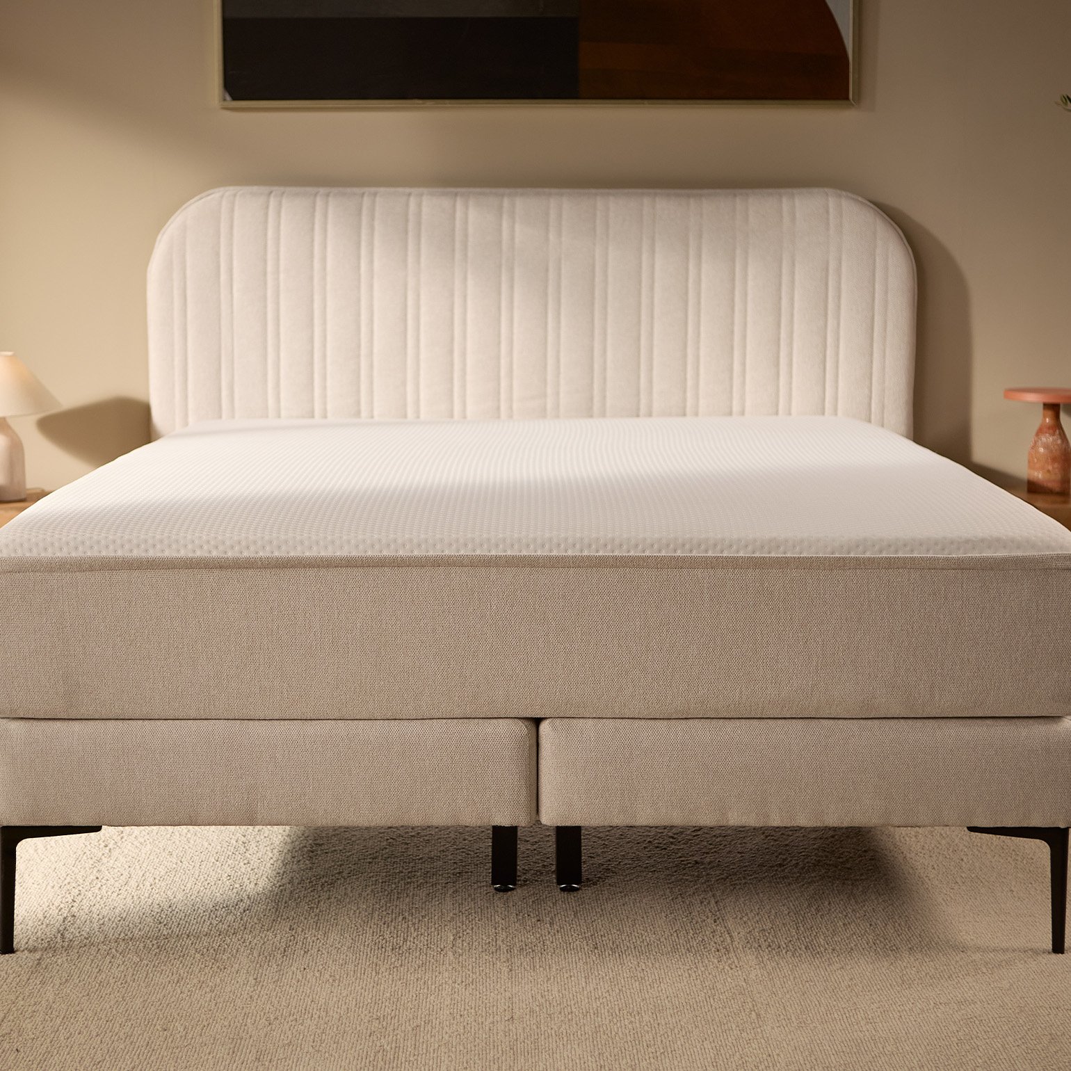 Frontansicht des kompletten beigen Bettes mit gestepptem Kopfteil, erhöhtem Boxspring-Unterbau, schlanken Außenbeinen und robusteren mittigen Stützbeinen.