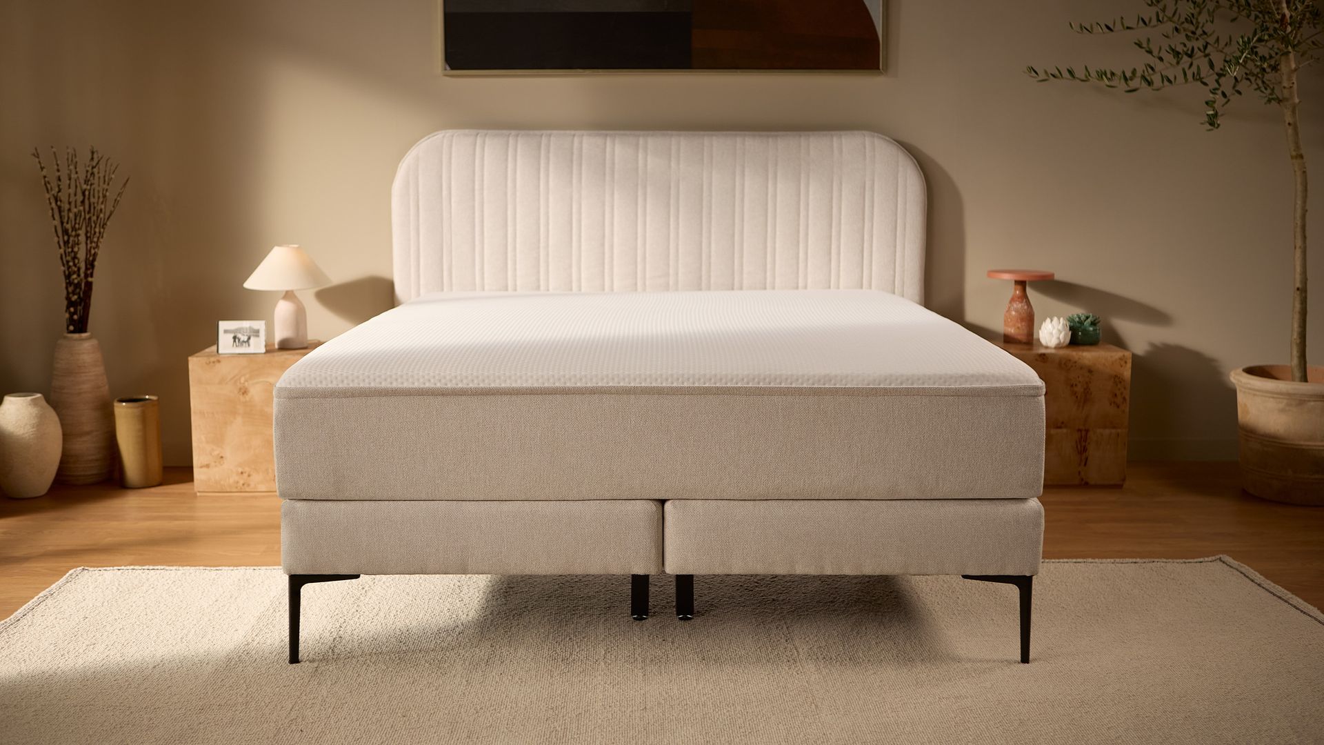 Frontansicht des kompletten beigen Bettes mit gestepptem Kopfteil, erhöhtem Boxspring-Unterbau, schlanken Außenbeinen und robusteren mittigen Stützbeinen.
