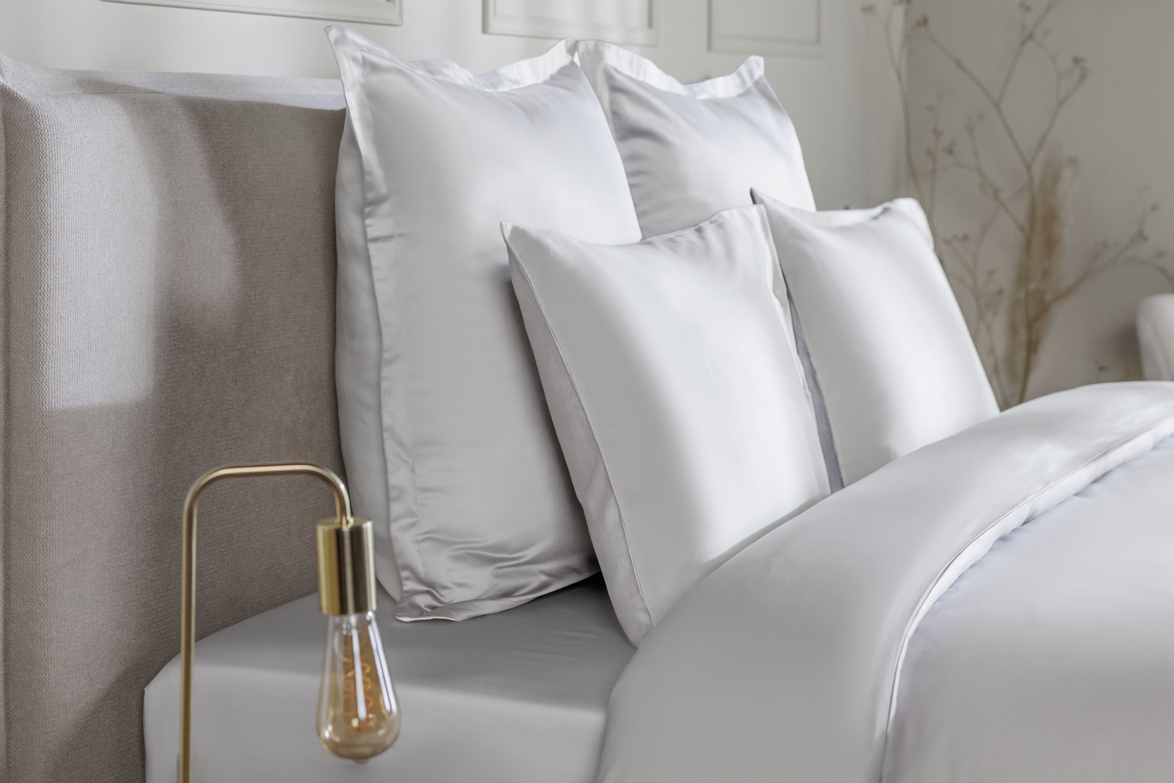Detail van een bedhoek met een Emma Satijn Hoeslaken in een tweekleurige combinatie van crème en beige, strak om een matras gespannen; hoeslaken heeft een subtiele glans en een zijdezachte afwerking die de luxe uitstraling benadrukt; bedframe is grijs met houten poten; de setting is minimalistisch met een witte muur, een lamp met gouden voet links, en droogbloemen aan de rechterzijde.