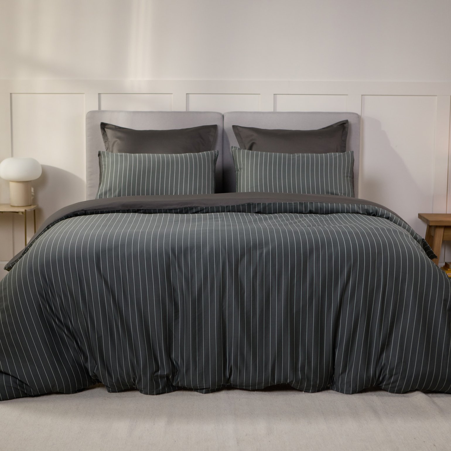 Slaapkamer met een opgemaakt bed in donkergrijs met strepen beddengoed, geflankeerd door twee houten nachtkastjes met lampen, planten en decoratieve manden.