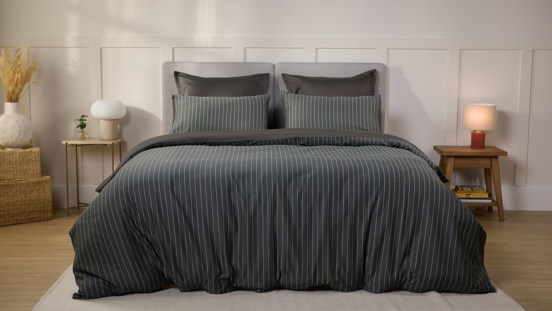 Slaapkamer met een opgemaakt bed in donkergrijs met strepen beddengoed, geflankeerd door twee houten nachtkastjes met lampen, planten en decoratieve manden.