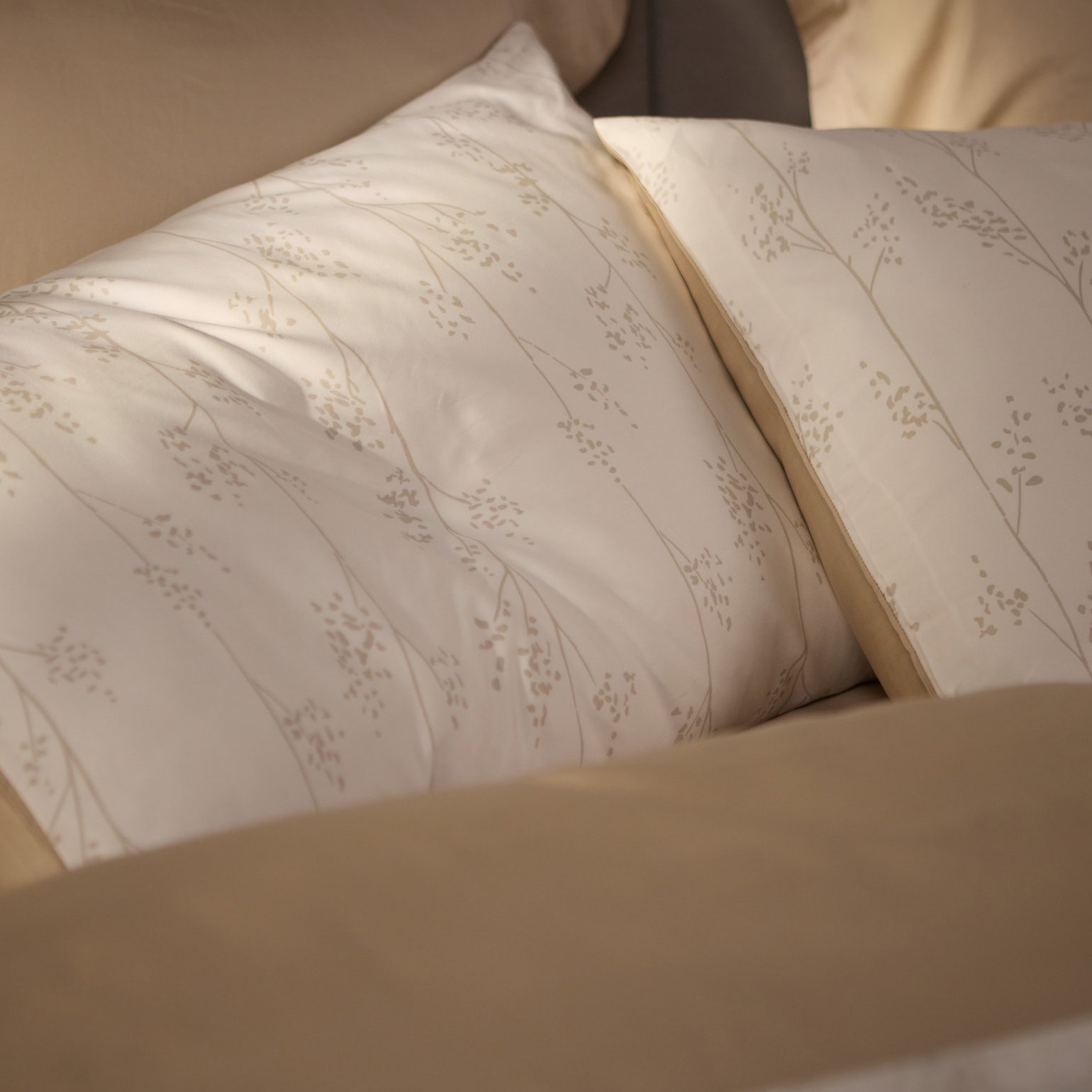 Close‑up van twee kussens met een subtiel botanisch patroon in lichttinten, omringd door beige beddengoed.