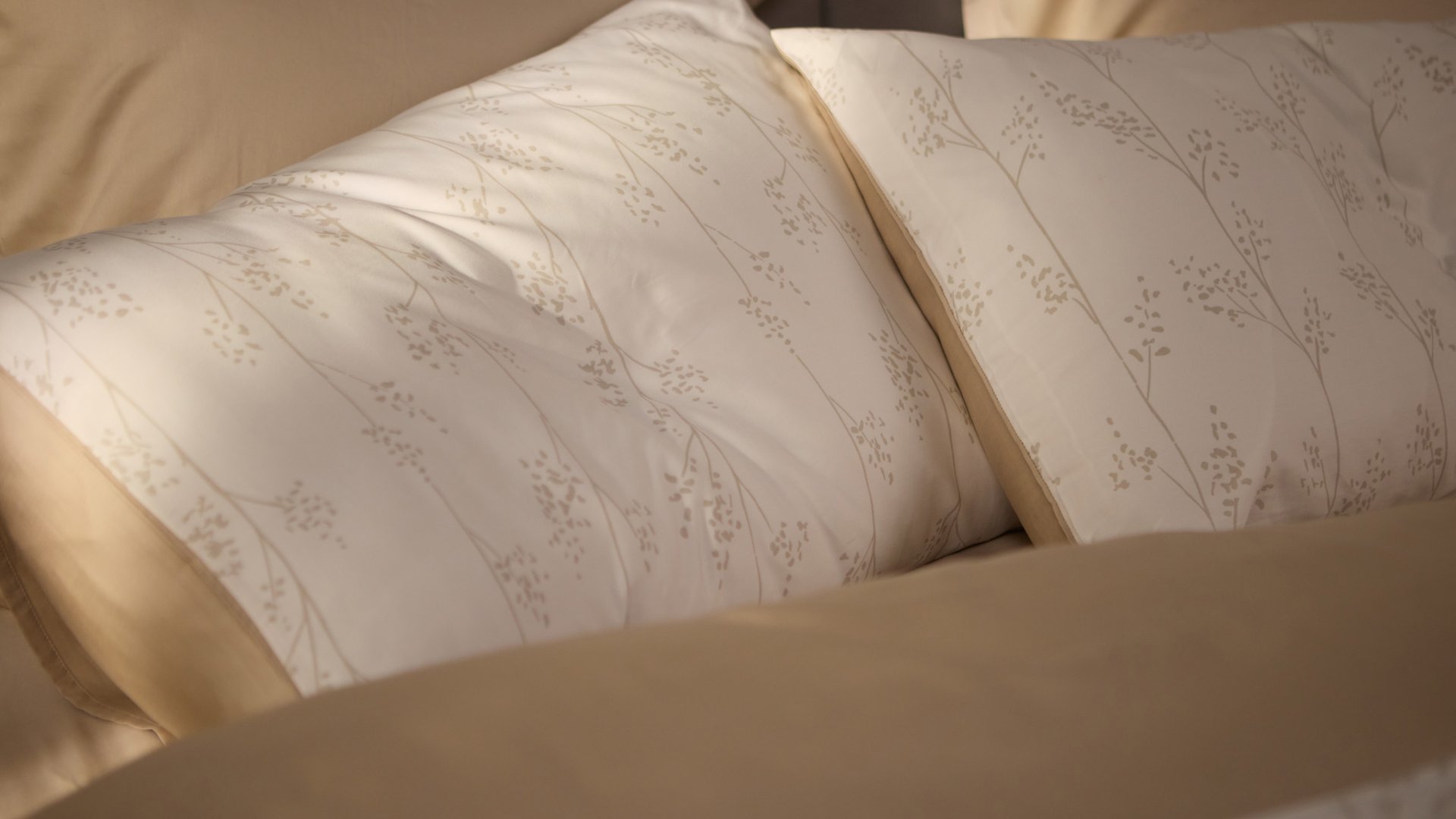 Close‑up van twee kussens met een subtiel botanisch patroon in lichttinten, omringd door beige beddengoed.