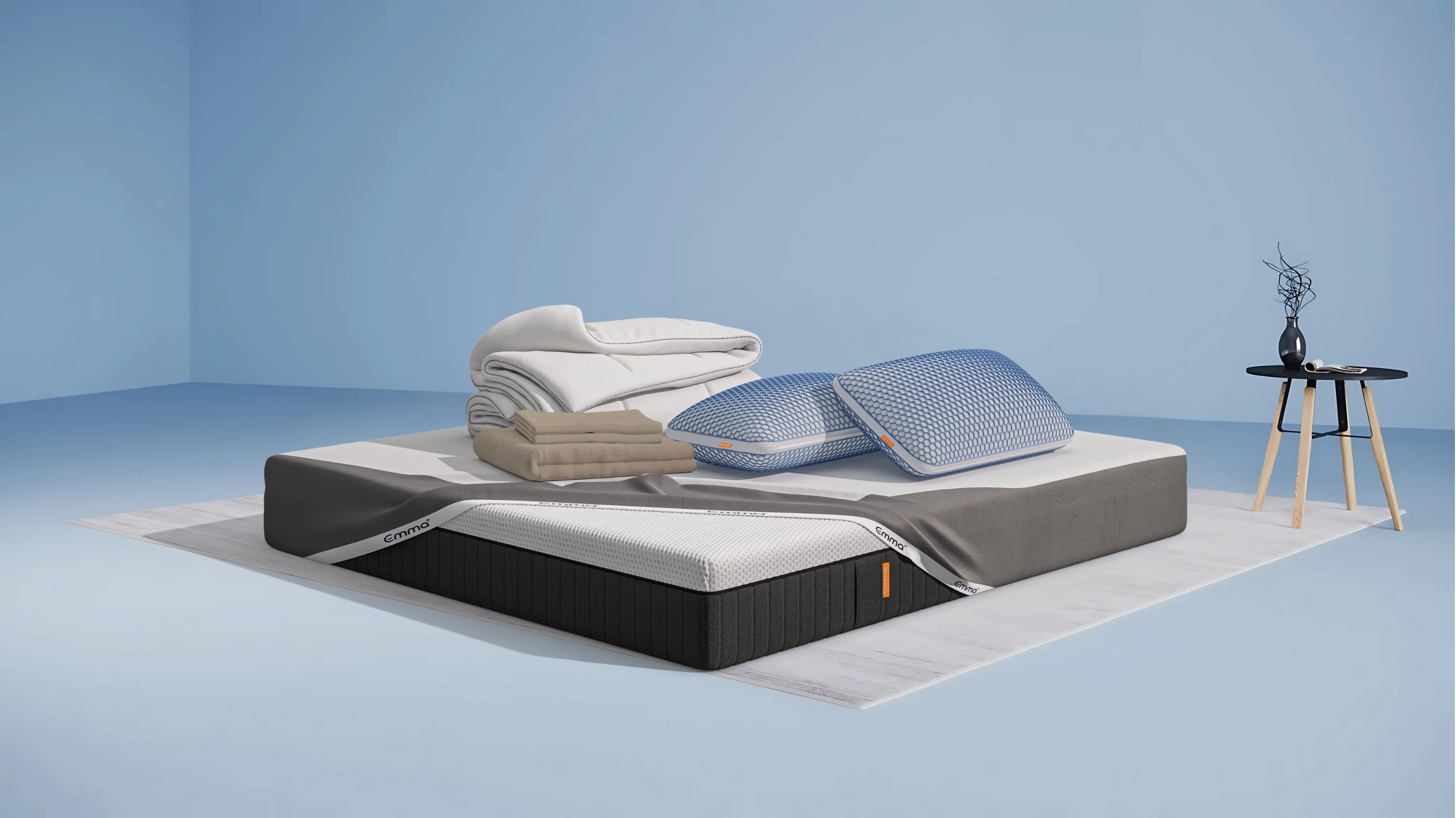 Ensemble de literie présenté dans une chambre moderne : matelas Hybride II ou Hybride Premium II avec base noire et surface blanche, partiellement recouvert d’un plaid gris foncé. Sur le matelas, deux oreillers Nuage Elite 40x70 cm bleus, une couette Nuage Tempérée blanche pliée, un protège-matelas, et une parure de lit en satin de coton blanc comprenant une housse de couette et des taies d’oreillers. Le tout est posé sur un tapis clair, avec une table d’appoint noire à pieds en bois et une décoration minimaliste.