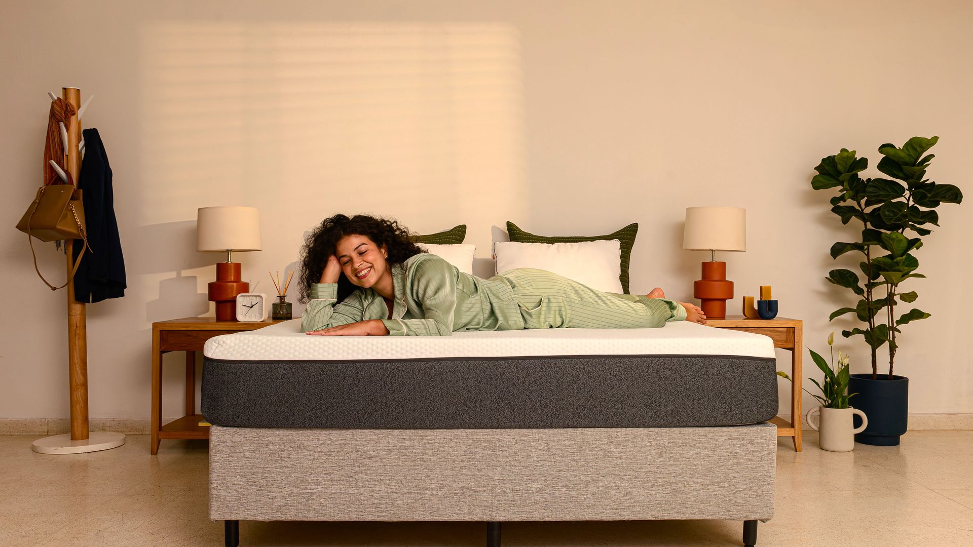 Quarto decorado com estilo minimalista e acolhedor. Há uma cama box com colchão Original Emma, cinza e branco, travesseiros brancos e uma moça deitada sobre ele, com pijama verde. Ao lado da cama, mesas de cabeceira com candeeiros e objetos decorativos. Um cabide com chapéu e casaco está à esquerda, e uma planta em vaso à direita