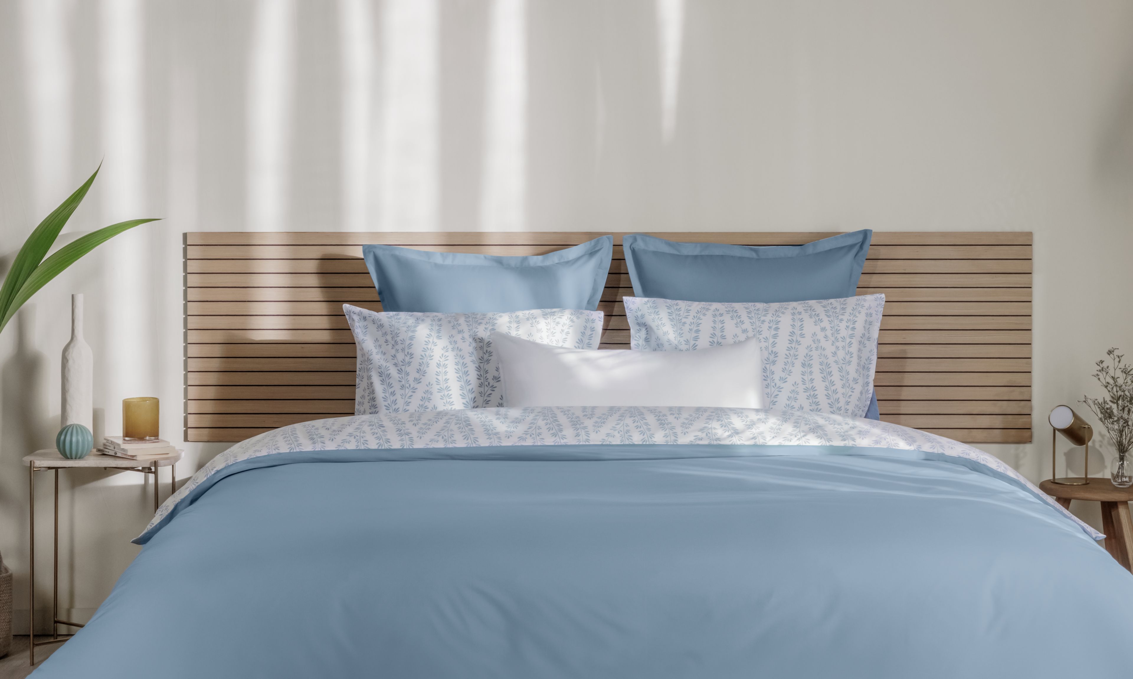 Letto ordinato con biancheria azzurro chiaro e bianca, testiera in legno e decorazioni naturali ai lati.