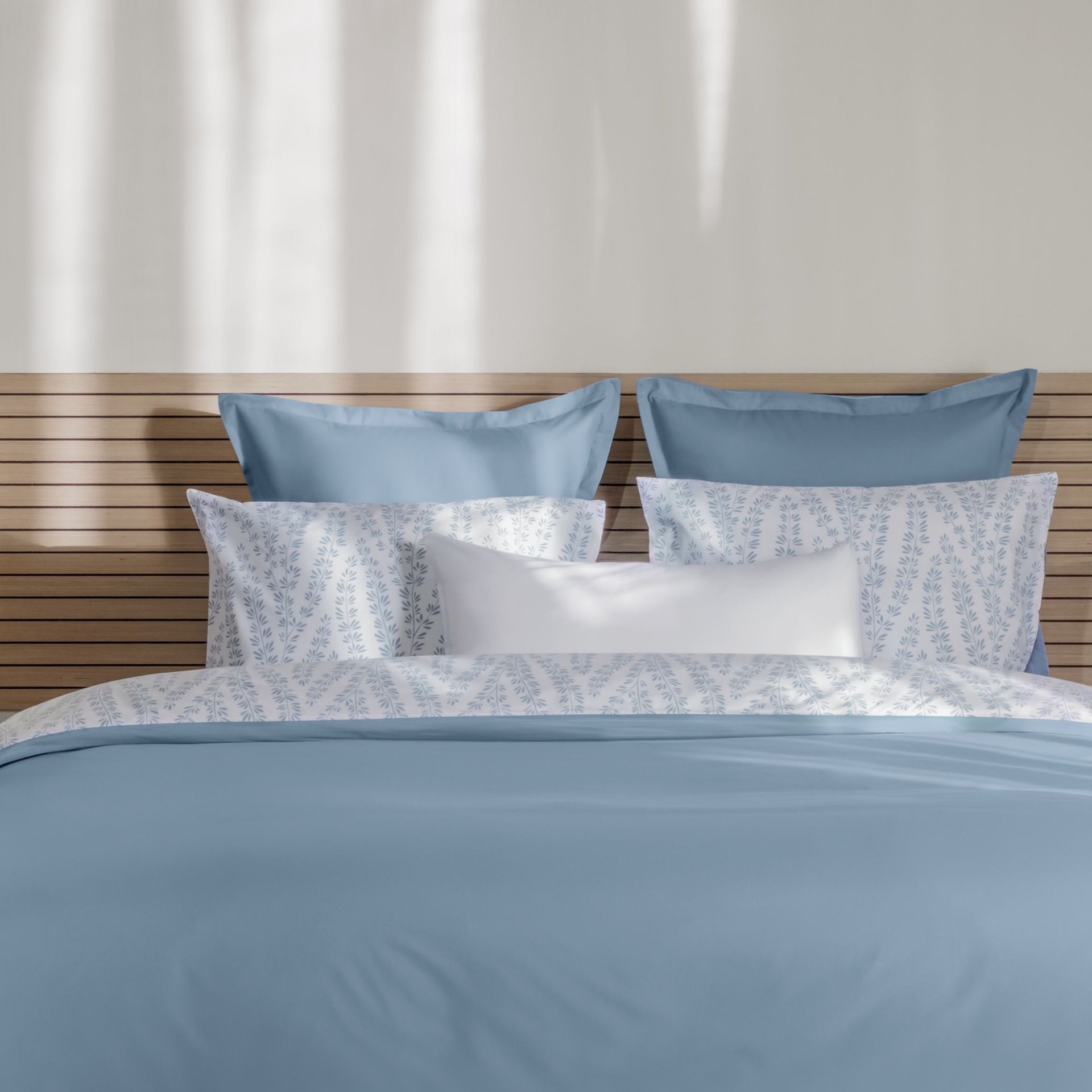 Letto ordinato con biancheria azzurro chiaro e bianca, testiera in legno e decorazioni naturali ai lati.