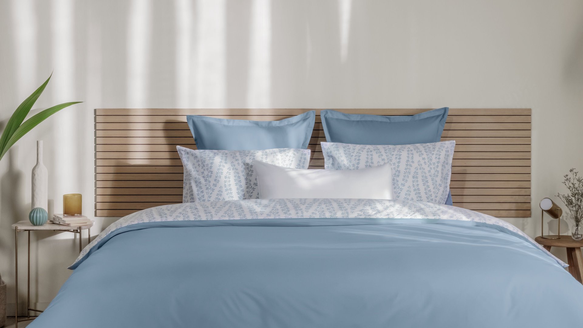 Letto ordinato con biancheria azzurro chiaro e bianca, testiera in legno e decorazioni naturali ai lati.